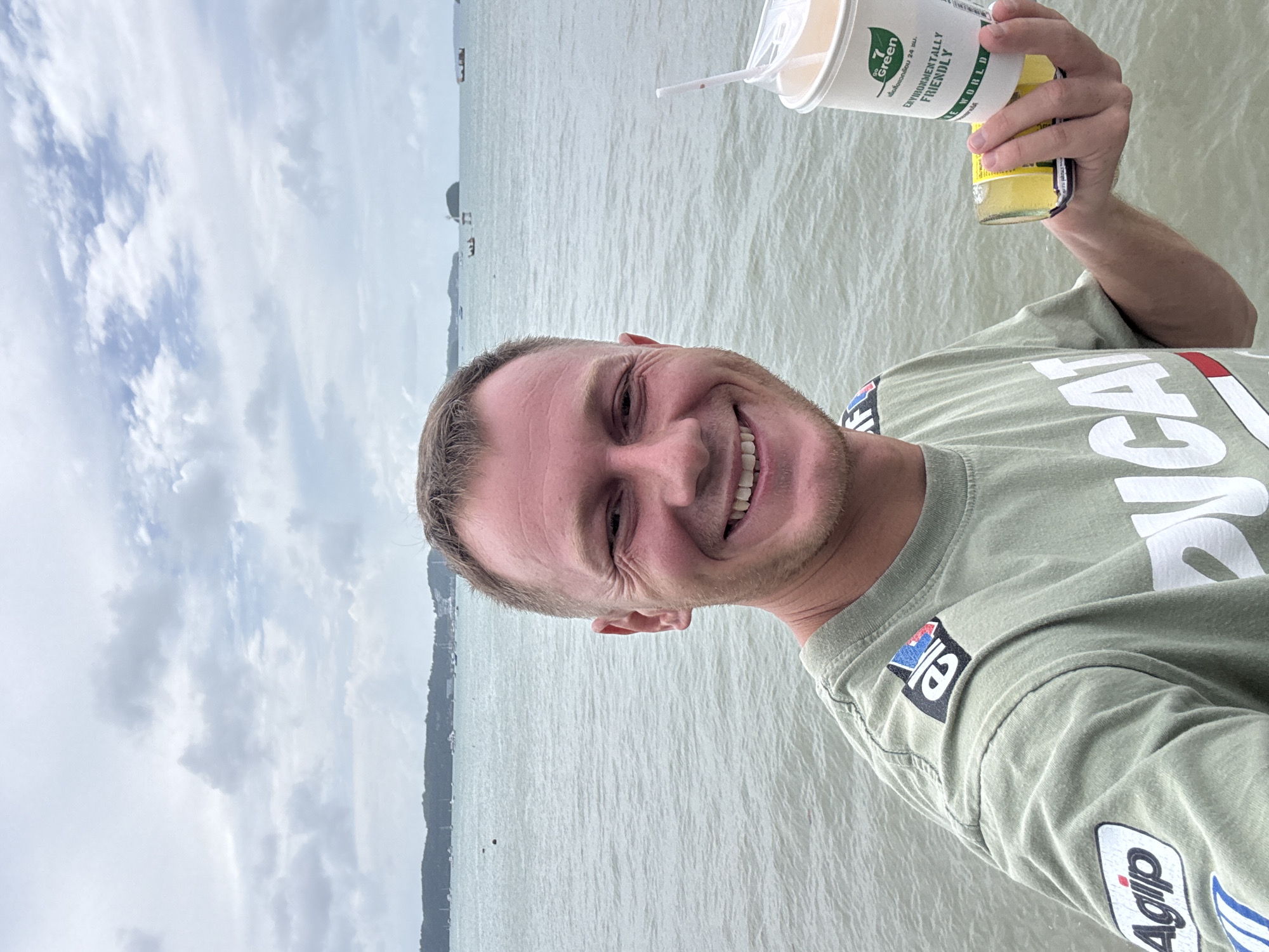 A man with short blond hair and a stubble is taking a selfie by the sea in Rawai, Thailand. He is wearing a light green t-shirt with the "Agip" and "Elf" logos on the left sleeve and shoulder. He is holding a paper cup with a straw in his right hand and a yellow can in his left hand. The sea is calm with a few boats in the background. The sky is cloudy. The man is smiling at the camera.