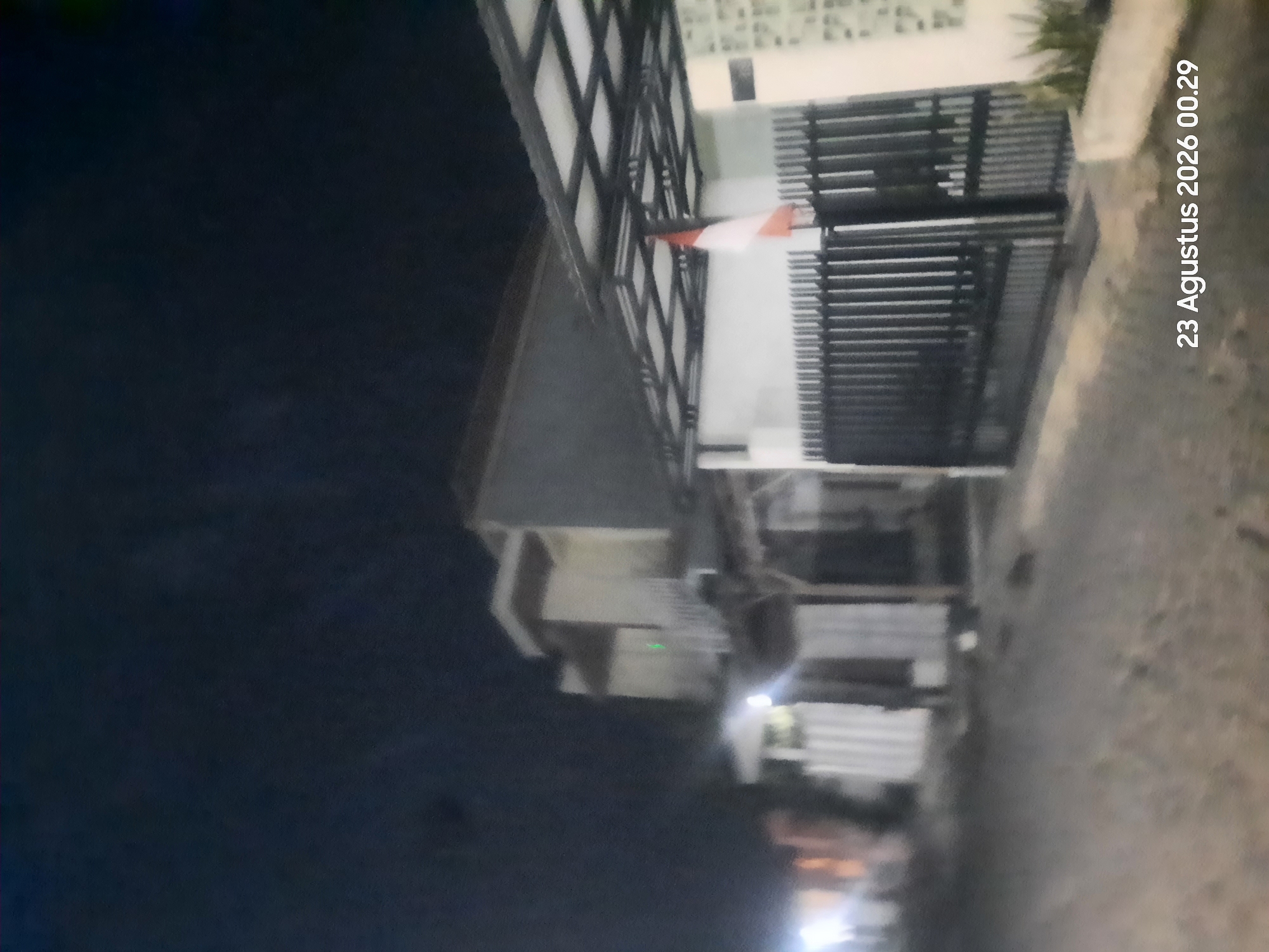 The image is a nighttime outdoor photograph of a residential street in Sukokerto, Indonesia. In the foreground, there is a dark, unpaved street with some debris. The middle ground features a black metal gate, partially obscuring a white-walled building with a red and white flag displayed. To the left, another building with illuminated windows is visible. The background is dominated by a dark, cloudless sky. The timestamp "23 Agustus 2026 00.29" is overlaid on the bottom right of the image.