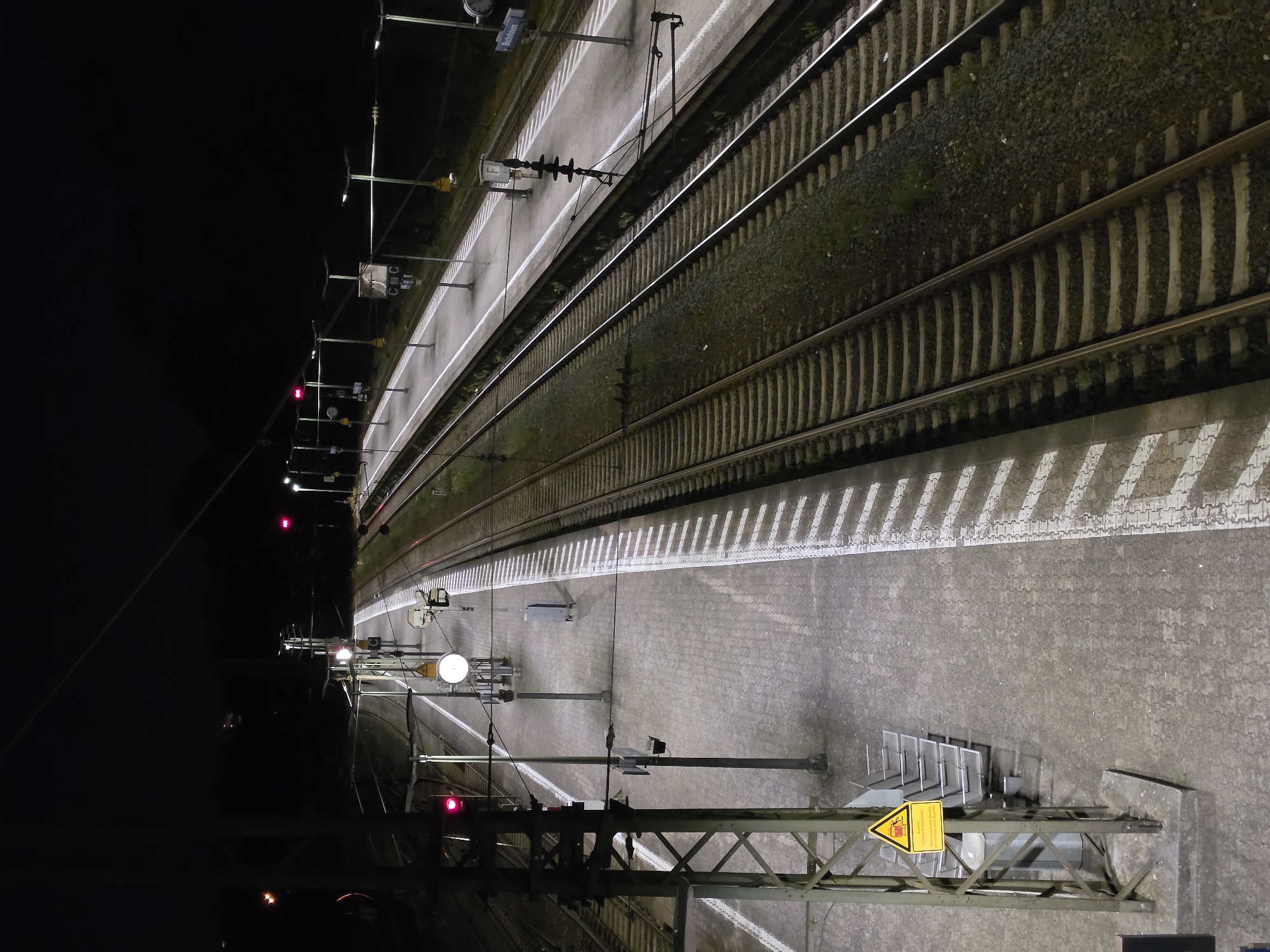 This is a nighttime, high-angle photograph of a train station in Bischofsheim, Germany. Multiple train tracks run parallel through the scene, bordered by paved platforms. Overhead electrical wires and supporting structures are visible. Lights illuminate the platforms and tracks. Several red signal lights are visible along the tracks, indicating train status. Benches are placed on the platform, and a trash receptacle is also present. A yellow warning sign with a pictogram of a falling object is mounted on a pole. The overall environment is dark, with artificial lighting creating shadows and highlighting the infrastructure.