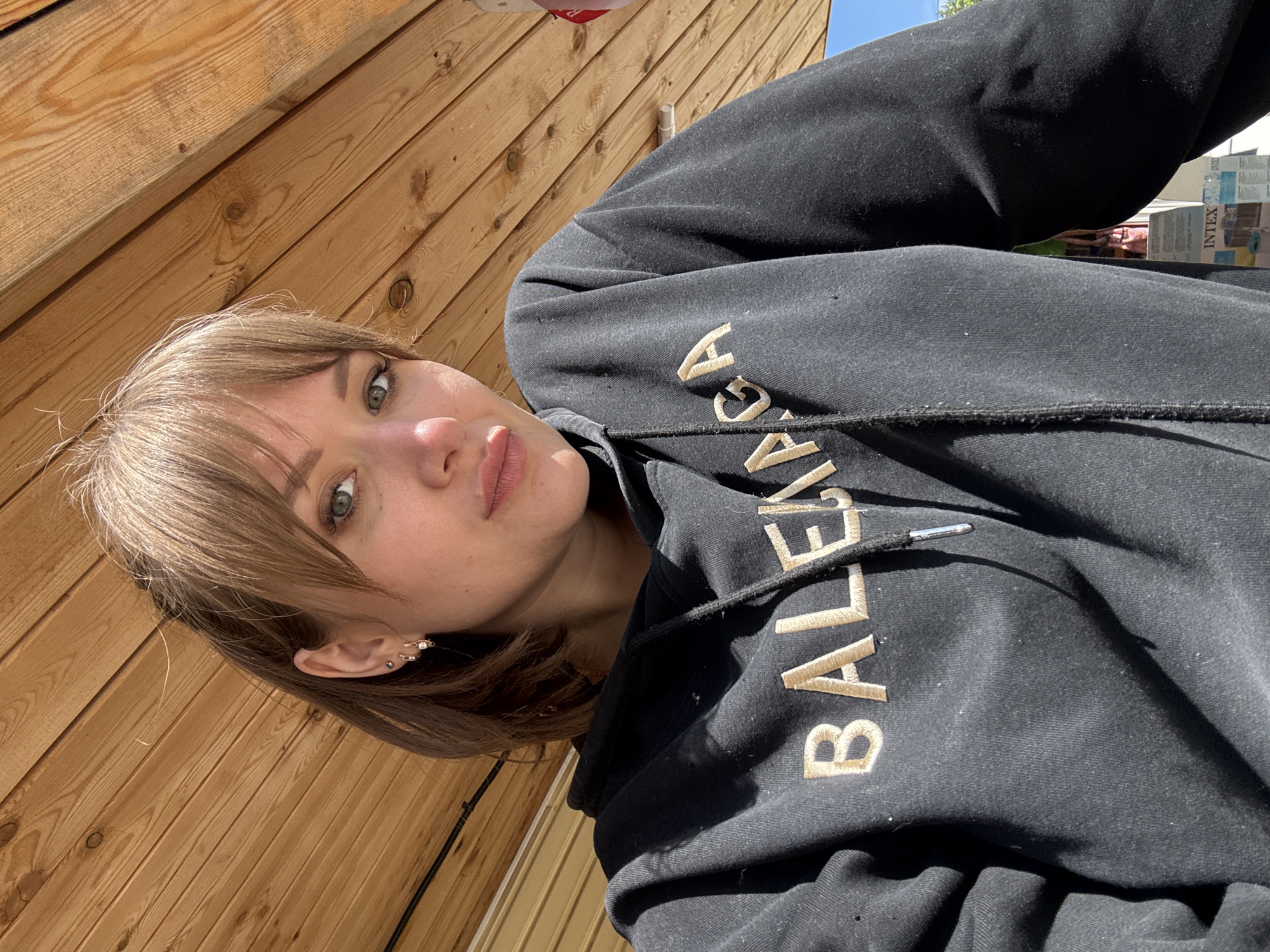 A light-skinned young woman with shoulder-length light brown hair and bangs looks directly at the camera, capturing a selfie from a low angle. Her green eyes are visible, and she wears light eye makeup and a light pink lip color, with her lips slightly pouted. Her left ear is adorned with three earrings: a small dark stud in the upper lobe, a small gold hoop with a dangling charm in the middle lobe, and another small gold hoop in the lower lobe. She is wearing a black hooded sweatshirt with "BALENCIAGA" embroidered in gold lettering across the chest. The sweatshirt fabric exhibits some wear or small fibers. The hood features a drawstring with silver-toned tips. The background overhead is a wooden slatted ceiling or roof structure, composed of light brown planks with visible grain and knots. To the lower right, a segment of clear blue sky is visible, along with a white structure and a partially visible cardboard box bearing "INTEX" branding multiple times. The scene is illuminated by bright, direct sunlight, creating strong highlights and shadows. This scene is located in the city of Chaikovsky, Russia.