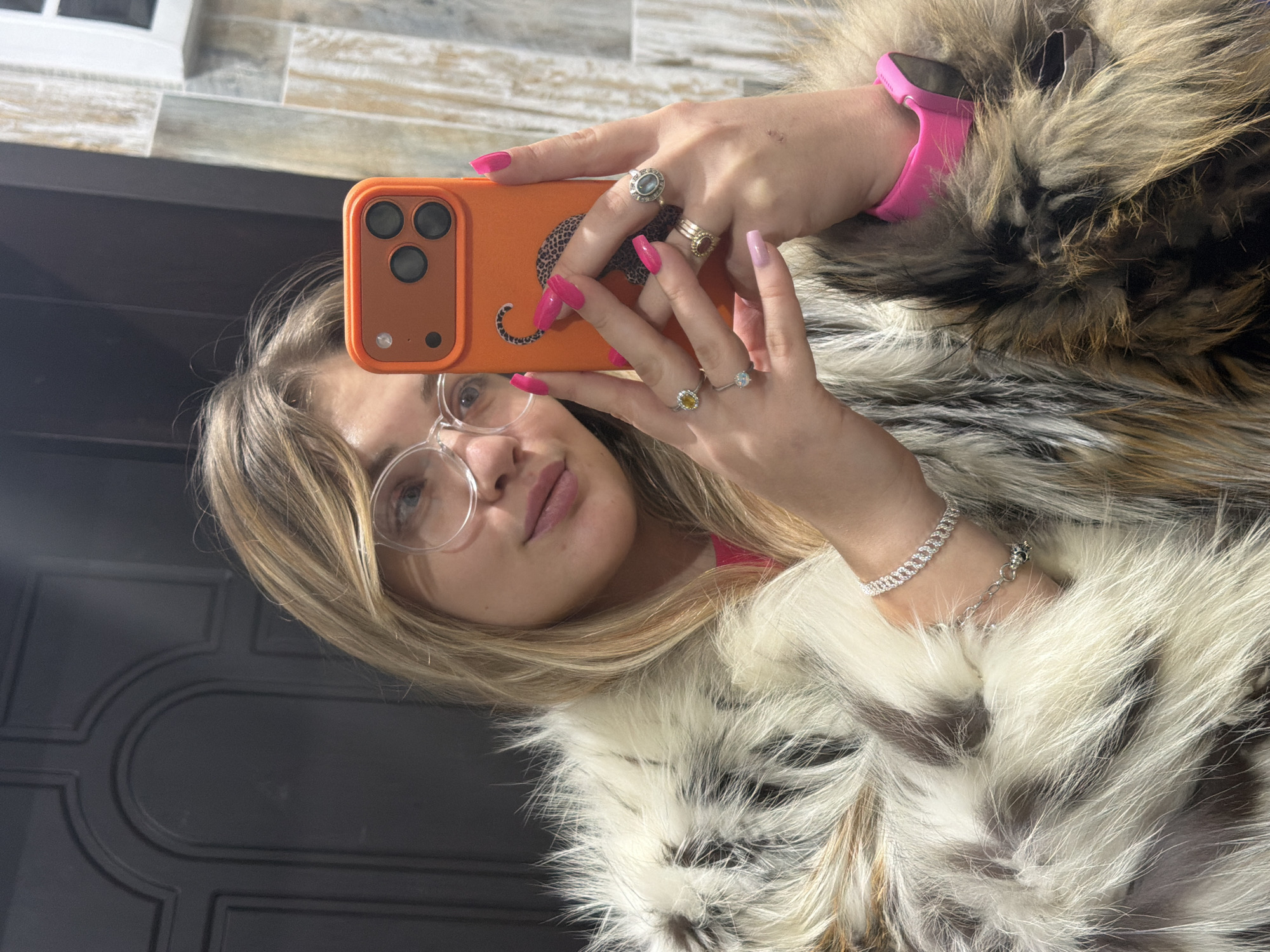 A young woman is captured in a mirror selfie, her reflection showing her face partially, featuring blonde hair, clear-rimmed glasses, and pink lips. She holds an orange smartphone, which has a black cheetah silhouette sticker on its back, obscuring part of her face. Her hands are adorned with several rings on multiple fingers, and her nails are painted bright pink on one hand and a lighter pink on the other. A pink smartwatch is visible on her left wrist, while her right wrist displays two silver chain bracelets, one with a small charm. She is wearing a prominent furry garment, appearing to be a coat or vest, characterized by mixed shades of brown, black, and white/cream fur. The background reflected in the mirror consists of an upper section with light-colored rectangular tiles and a lower section of a dark, possibly paneled, surface or door. The scene is indoors, located in Большая Шильна, Russia. No visible text is present in the image.