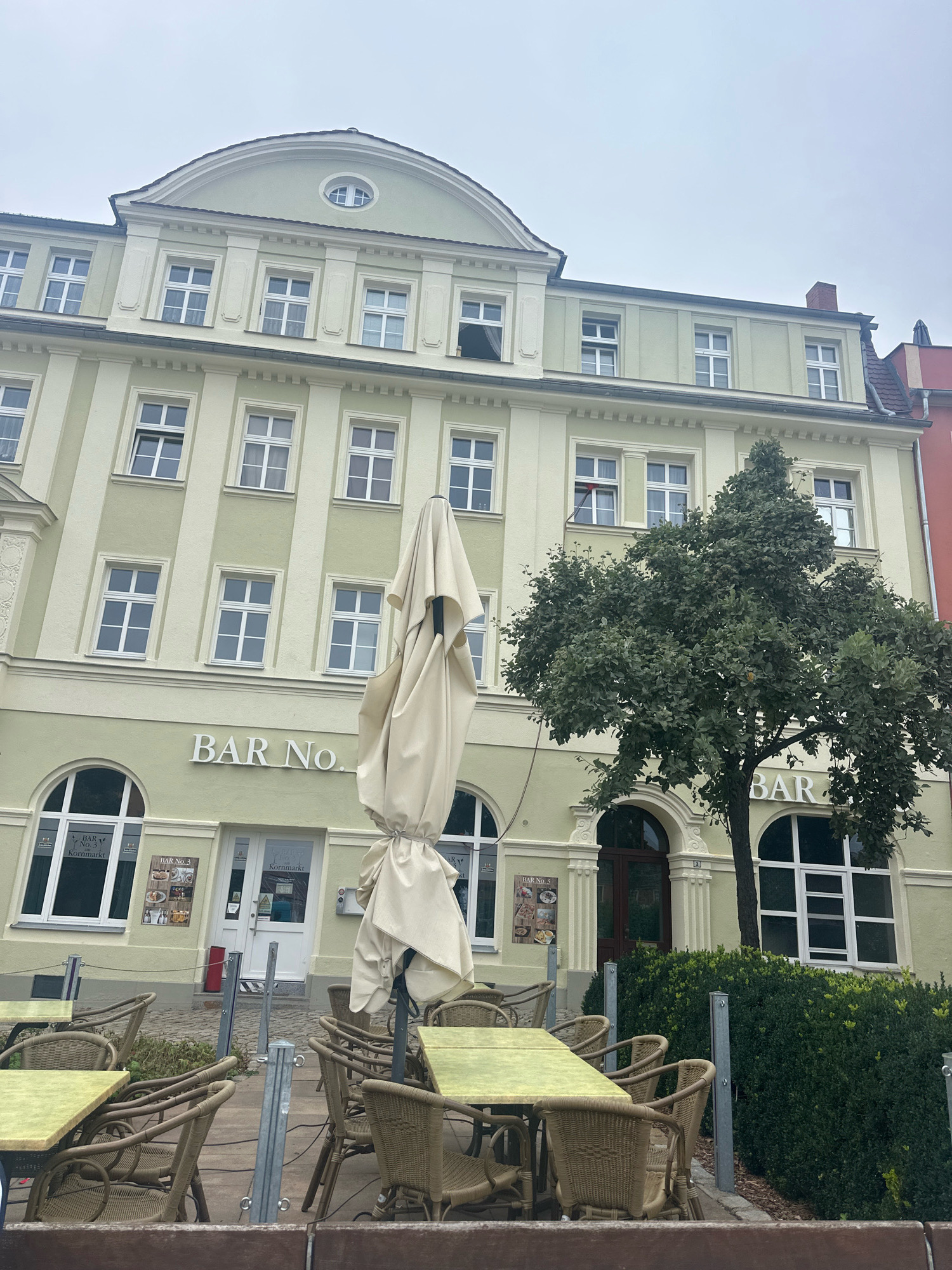 The image shows the exterior of a building in Bautzen, Germany, with an outdoor seating area for a bar or restaurant. The building is light green with white trim and has multiple windows on its upper floors. The ground floor features a storefront with the words "BAR No." prominently displayed. Several tables and chairs made of wicker are set up outside, along with a large, folded umbrella. A tree with green foliage stands to the right of the building. The sky is overcast. The scene appears to be a quiet street with no people visible.