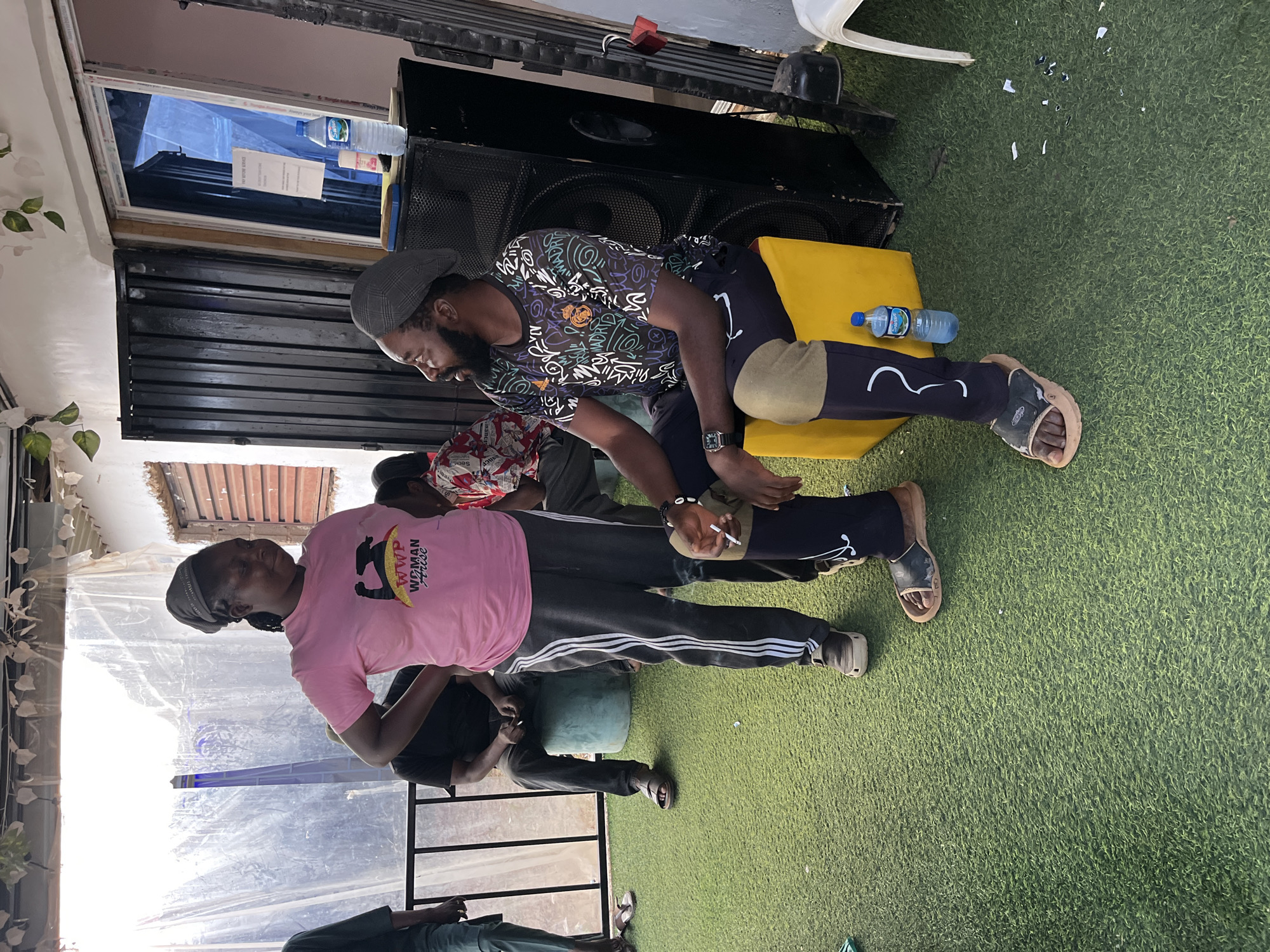 In a vaguely defined outdoor urban setting, possibly a courtyard or patio area with artificial turf flooring, two individuals are engaged in conversation. The woman, wearing a pink t-shirt and dark grey sweatpants with white stripes, stands while looking at the seated man. The man, dressed in a black t-shirt with a colorful graphic print and dark pants, is seated on a yellow cube, facing the woman. He wears a grey cap and has a beard. Both individuals appear to be holding cigarettes, with smoke visible. Behind them, a large black speaker system is positioned next to a building with metal shutters and a window. A blue water bottle rests on the ground near the yellow cube. Other individuals are present in the background, some seated and others standing, and some also appear to be smoking. The environment is partially covered by a translucent tarp overhead.