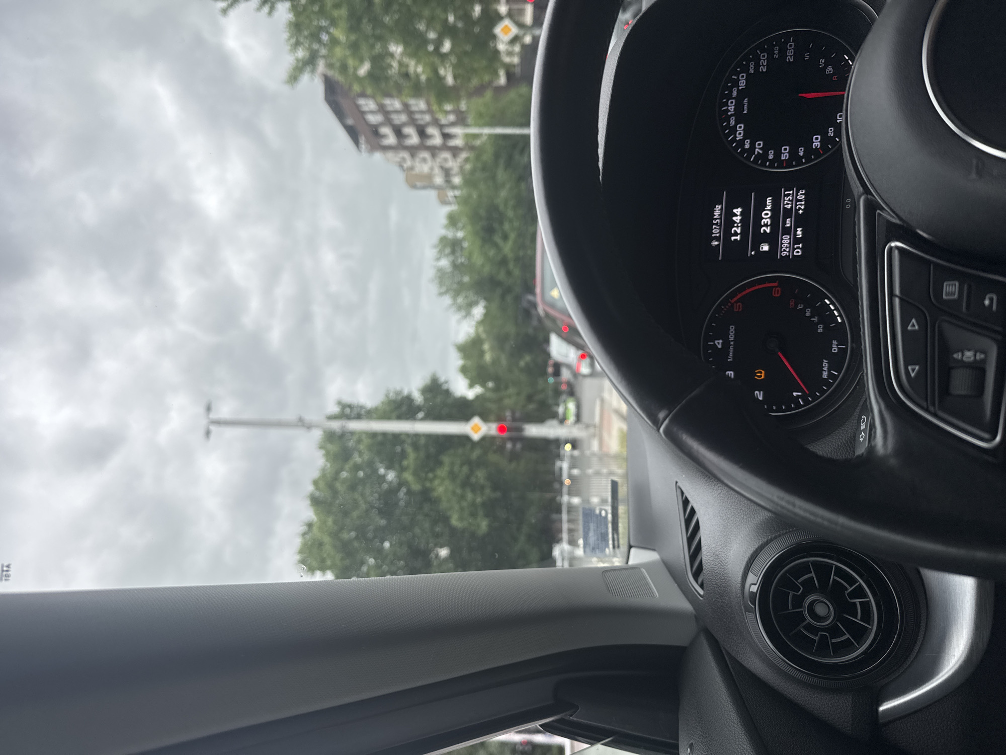 The image provides a view from inside a car, looking forward and slightly to the left, capturing both the vehicle's interior and an urban street scene under an overcast sky.
In the foreground and to the right, a black steering wheel partially obscures the instrument cluster. The visible dashboard displays essential information: the time is "12:44", the car's odometer reads "92980 km", and the trip meter shows "475.1 km". The outside temperature is "+21.0°C", and the remaining fuel range is "230 km". The radio is tuned to "107.5 MHz". The gear indicator shows "D1", and both the speedometer and tachometer display zero, indicating the car is stationary. Other text visible on the dashboard includes "km/h", "1/min x 1000", "READY", "OFF", and "LIM". A round, black air vent is visible to the lower right of the steering wheel.
Outside the car, an urban environment is visible through the windshield. The car is stopped at a red traffic light, clearly visible in the midground. Beyond the traffic light, other vehicles are present on the street. Lush green trees line the street, partially obscuring a multi-story building with many windows in the background on the left. The sky is completely overcast, suggesting cloudy weather. This scene is located in Plovdiv, Bulgaria.