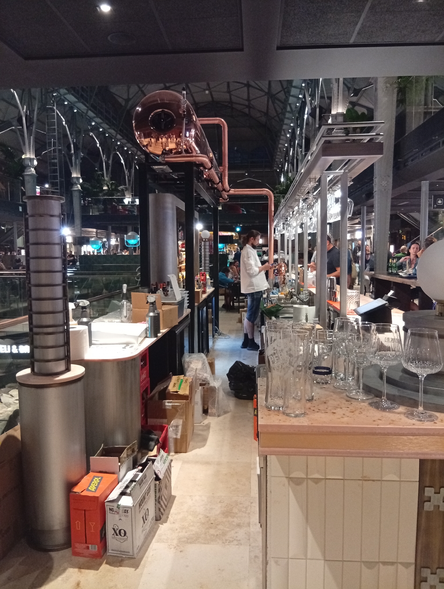 The image depicts an indoor scene, likely a bar or a food court, possibly located in the city of Gdańsk, Poland, given the presence of branded items that may be associated with the region. In the foreground, a bar counter is visible, adorned with a variety of glassware, including pint glasses and wine glasses. Behind the counter, a person in a white chef's jacket and blue shorts stands, appearing to be engaged in work. The background reveals a multi-level structure with exposed metal beams and ceiling, illuminated by numerous lights. Other individuals are present in the background, some seated at tables, suggesting a public gathering space. The overall ambiance is that of a modern, well-lit establishment.