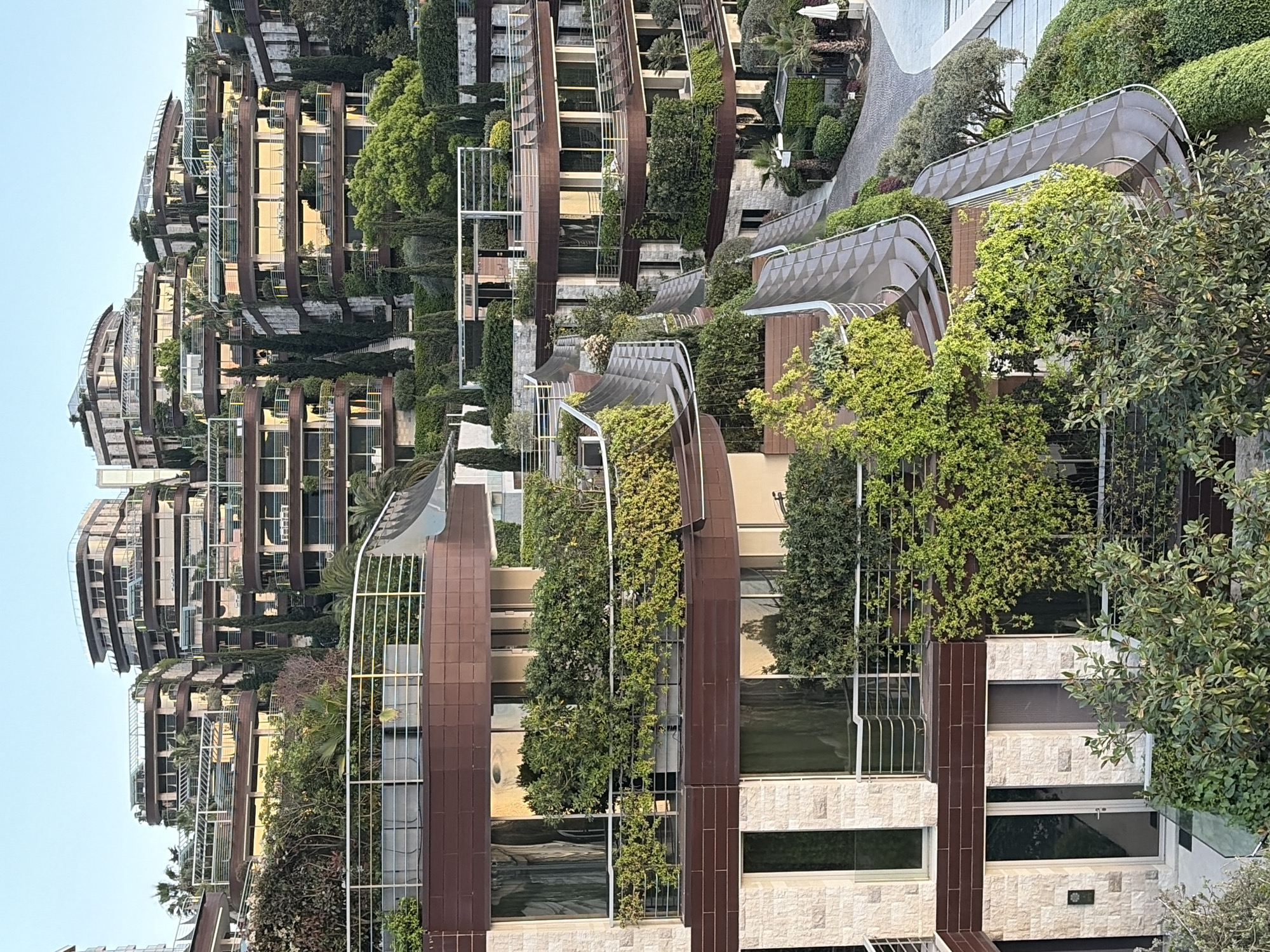 The image depicts a multi-story residential building in Budva, Montenegro, characterized by its curved balconies and abundant vegetation. The architecture features a combination of earth-toned materials, including brown paneling and light-colored stone. Each balcony is lined with a variety of plants and trees, creating a "vertical garden" effect. The building is situated in an area with lush greenery, including mature trees and manicured gardens. Overhead structures with curved, semi-transparent roofs are visible, suggesting outdoor walkways or seating areas. The sky appears pale blue, indicating daylight. The overall impression is of a luxurious, eco-friendly residence integrated into its natural environment.