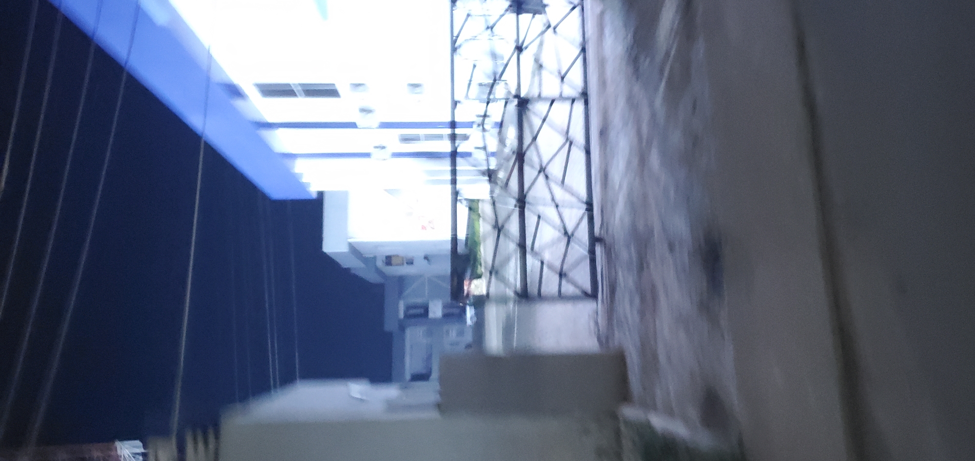 This image, captured at night or in very low light conditions within Jos, Nigeria, displays significant motion blur across the entire frame. The scene is viewed from a low perspective, likely from a paved ground level.
In the foreground, a dark-colored metal gate with a criss-cross or triangular geometric pattern spans horizontally, appearing closed. To the immediate left of the gate, a thick, light-colored structural element, possibly a column or wall, is partially visible. The ground directly in front of the gate and column is a dark, uneven surface, consistent with a paved area such as a road or courtyard, exhibiting streaks and variations in tone due to the low light and blur.
Beyond the gate, several buildings are visible. On the right, a prominent multi-story building with white walls and a distinct blue upper band or trim is brightly illuminated, appearing overexposed in areas due to intense light sources from within or nearby. This building features multiple rectangular windows and possibly air conditioning units mounted on its exterior. Further into the mid-ground and to the left, additional light-colored, multi-story buildings with flat roofs are discernible, though less distinctly due to distance and blur. One structure in the mid-left background has a darker, possibly grey, upper section.
The sky occupies the upper portion of the image, appearing as a dark indigo or deep blue hue. Against this sky, several thin, parallel lines, consistent with overhead power lines or communication cables, stretch horizontally across the frame.
No individuals, specific actions, or interactions are discernible within the image. The environment suggests an urban or semi-urban institutional or residential complex, characterized by modern architecture and a secure entrance.