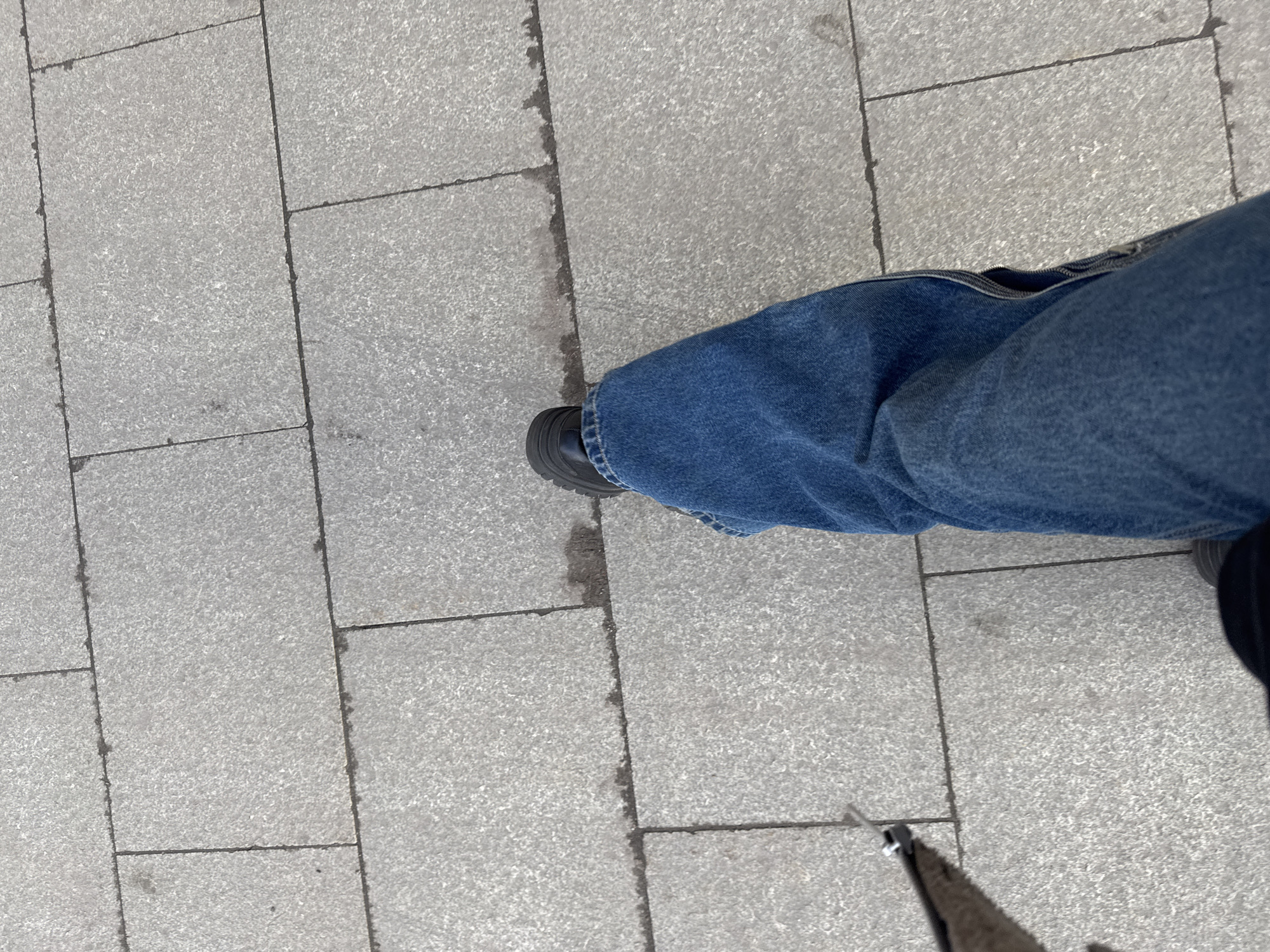 The image shows a section of a paved sidewalk with a pair of legs wearing blue jeans and black shoes. The sidewalk is composed of rectangular gray paving stones. The legs are positioned towards the right side of the frame. The city of Moscow, Russia is not discernible from the information available.