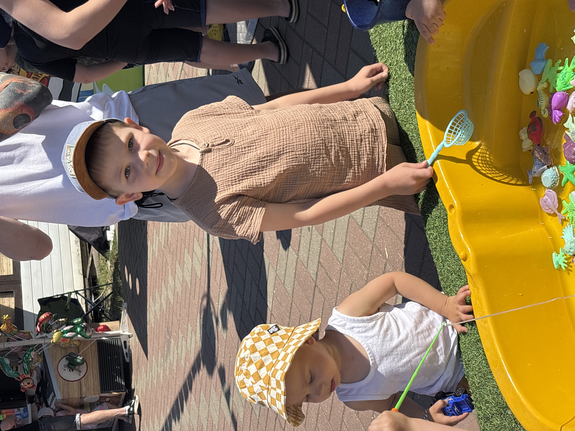 Two young boys are playing in a yellow plastic wading pool filled with water and small plastic sea creatures. The boy on the right, wearing a tan shirt and a baseball cap, is looking towards the camera with a slight smile. The boy on the left, wearing a white tank top and a checkered bucket hat, is holding a blue toy truck and a green fishing pole. The wading pool is placed on a patch of artificial grass, and behind them is a brick pathway. In the background, a white building and a stand with colorful balloons are visible. The scene appears to be outdoors on a sunny day. The location is identified as Rusanovo, Russia.