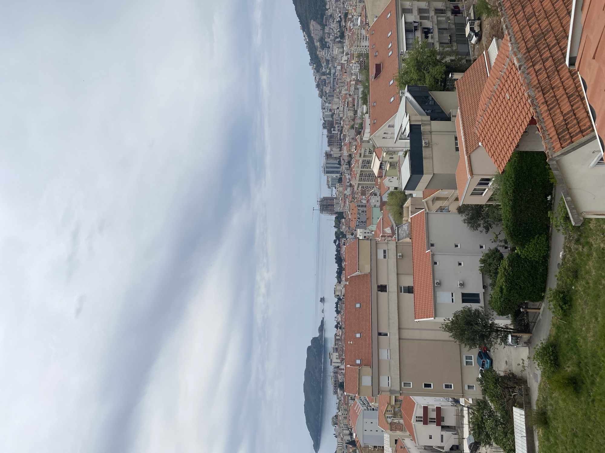 The image shows a view of Budva, Montenegro. The foreground features buildings with red tile roofs and beige walls. Trees and greenery are interspersed among the buildings. In the background, the Adriatic Sea stretches between the mainland and Sveti Nikola Island. Buildings line the coast along the sea. The sky is overcast with light-colored clouds.