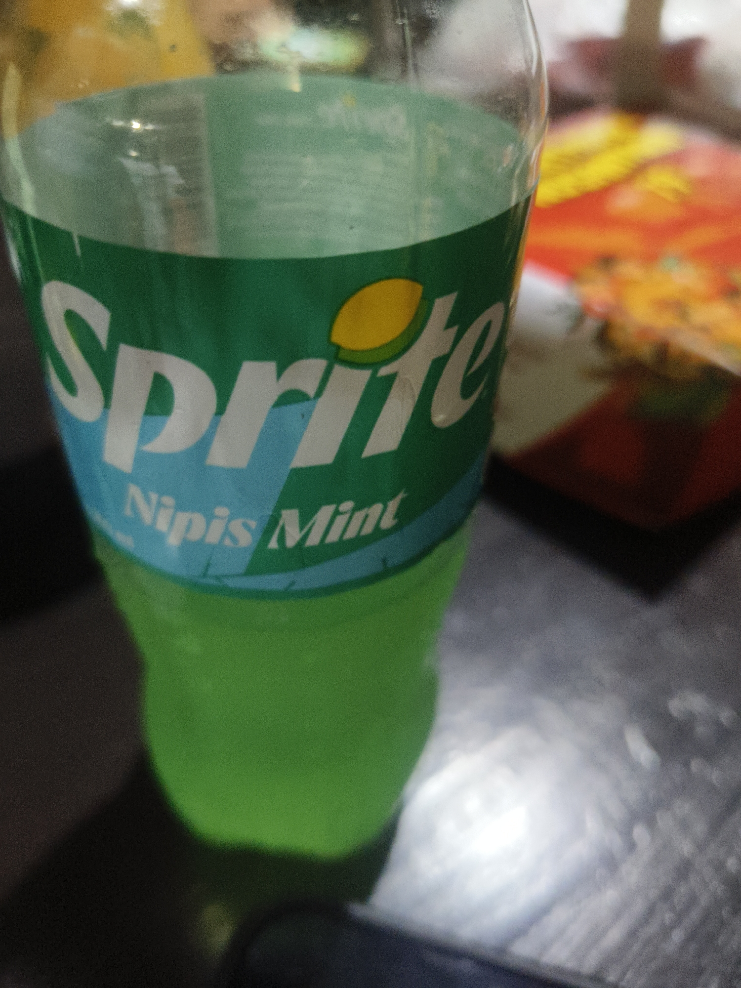 The image shows a close-up of a Sprite bottle with a green label and white lettering. The word "Sprite" is prominently displayed in large white letters, with a yellow lemon graphic to the right. Below "Sprite," smaller white text reads "Nipis Mint." The bottle is filled with a green liquid. The background is out of focus, showing a blurred colorful surface, possibly a table with some items on it, and a darker area to the left which could be a shadow. There are no people visible in the image, and no specific landmarks that would indicate a location in Kuta Selatan, Indonesia. The scene appears to be an indoor setting, possibly a home or a restaurant.