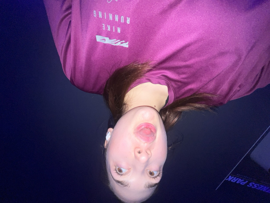 A woman is seen wearing a purple Nike running shirt. She has brown hair and light skin. Her mouth is open and her tongue is visible. She's wearing a white wireless earbud in her left ear. The background is dark, appearing to be a dimly lit space. The words "FITNESS PARK" appear backwards on a screen in the lower right corner.