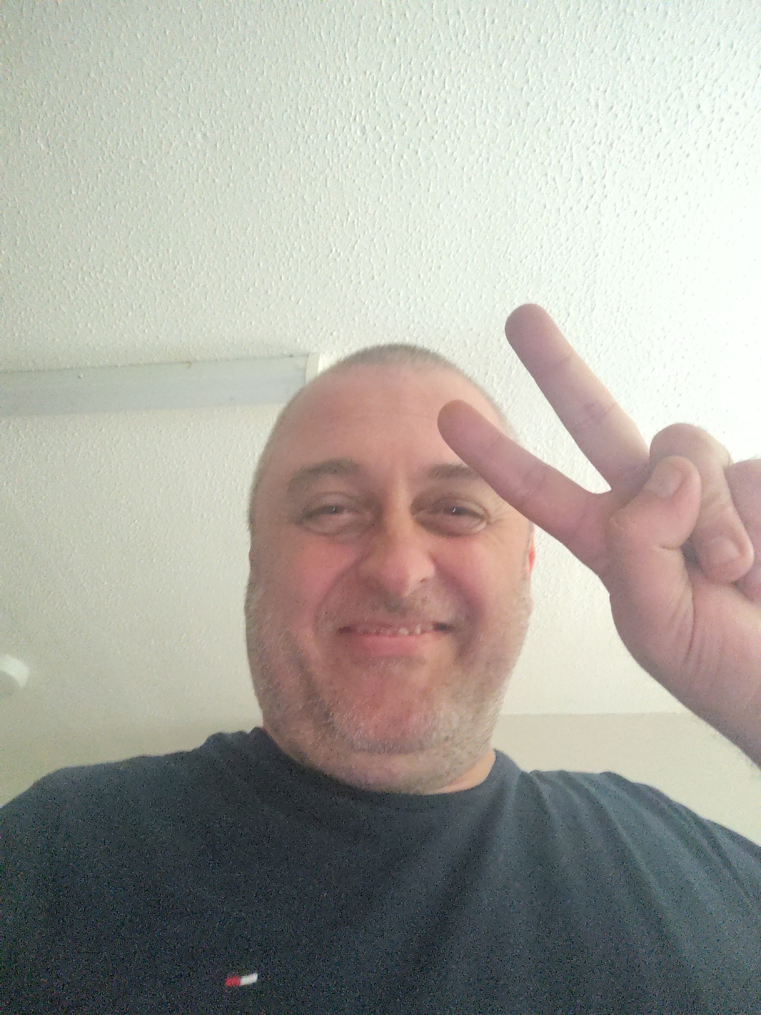 This image, captured in Huntingdonshire, United Kingdom, features a smiling, fair-skinned male, likely in his 40s or 50s, looking directly at the camera. He has short or bald hair on top and a short, greyish stubble beard. He is wearing a dark, short-sleeved t-shirt, which displays a small rectangular logo with horizontal red, white, and blue stripes on the lower left side (viewer's left). His right hand is prominently featured, held up to form a two-finger "peace sign" (V-sign) with his index and middle fingers extended. The background consists of a plain, off-white ceiling or wall with a distinct, rough "popcorn" or stucco-like texture. Visible in the upper left corner of the background is a portion of a white circular object, possibly a light fixture or smoke detector, and a horizontal white strip runs across the upper mid-section of the background. The perspective suggests a close-up selfie taken from a slightly low angle.