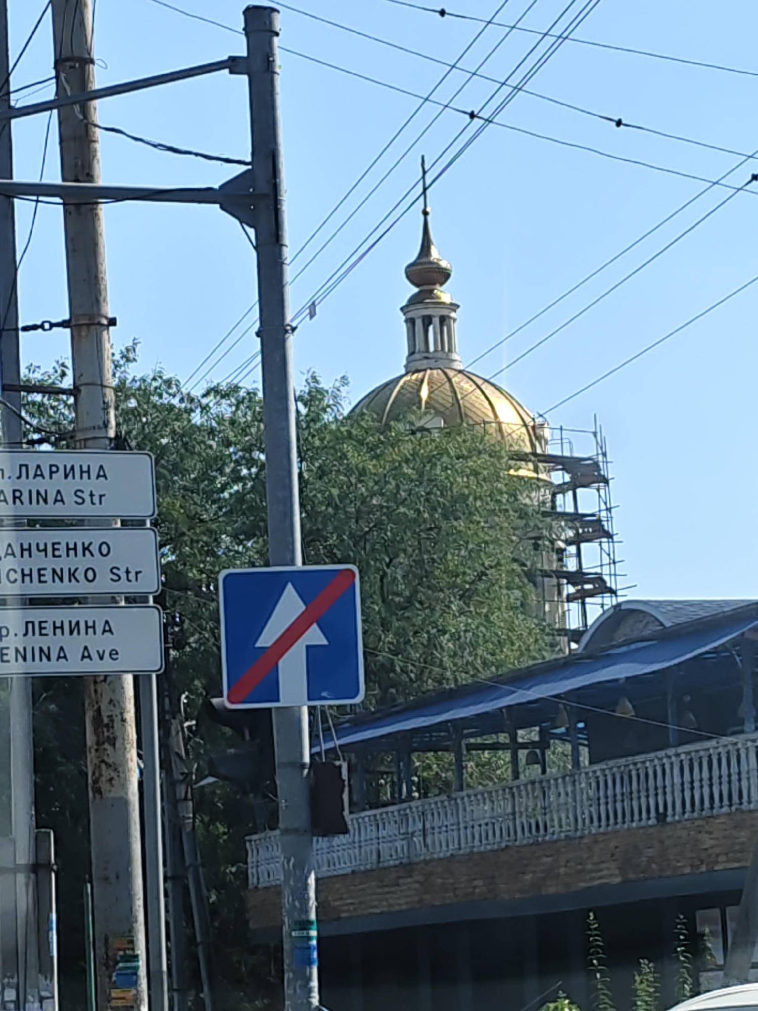 The image captures a street scene in Rostov-on-Don, Russia, on a clear day. Prominently featured are traffic signs and utility poles supporting a web of overhead wires. Three rectangular street signs are mounted vertically on a pole. The top sign reads "п. ЛАРИНА" and below it "ARINA Str". The middle sign displays "ДАНЧЕНКО" and "ICHENKO Str". The bottom sign says "р. ЛЕНИНА" and "ENINA Ave". To the right of these signs, a blue square traffic sign with a white upward arrow intersected by a red diagonal line indicates the end of a one-way street or lane. In the background, a golden dome of a building, possibly a church or administrative building, is visible against the blue sky, with scaffolding attached to its side, suggesting ongoing construction or renovation. Lush green trees partially obscure the lower portion of the dome. Below the dome and trees, a structure with a blue roof and a wooden balcony with decorative railings is visible. The overall scene depicts a typical urban environment with elements of infrastructure, signage, and architectural features.
