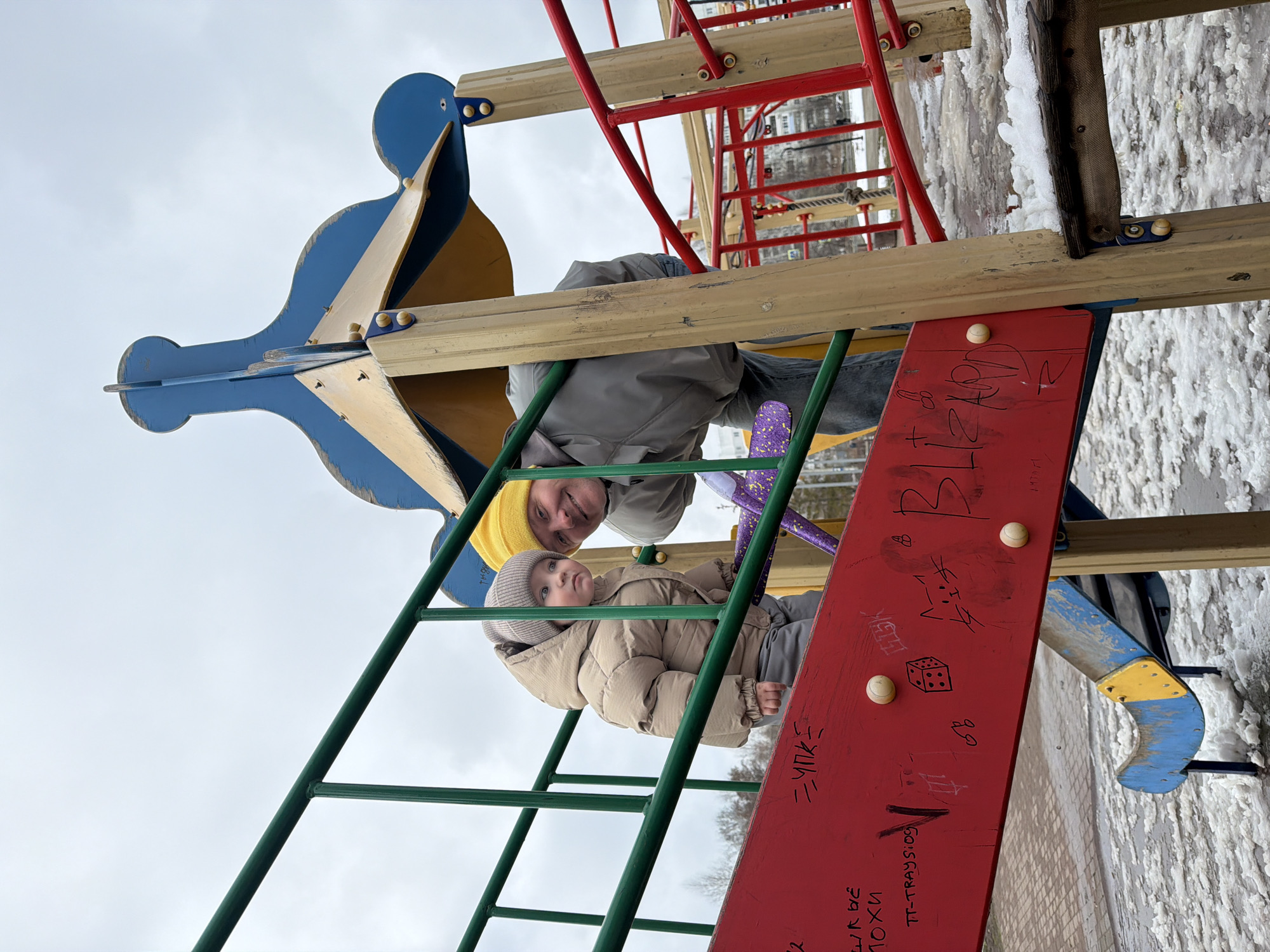 A man and a young child are positioned on a playground structure in Chaikovsky, Russia. The man, wearing a grey jacket and yellow beanie, looks down at the camera, his face partially obscured by the playground's wooden beams. The child, wearing a beige padded coat and grey hat, sits on a green metal ladder leading up to a red slide. The slide is marked with graffiti, including a drawing of a cat and a dice. The playground equipment is made of wood and metal and features a blue, gold, and red color scheme. Snow covers the ground. The sky above is overcast.
