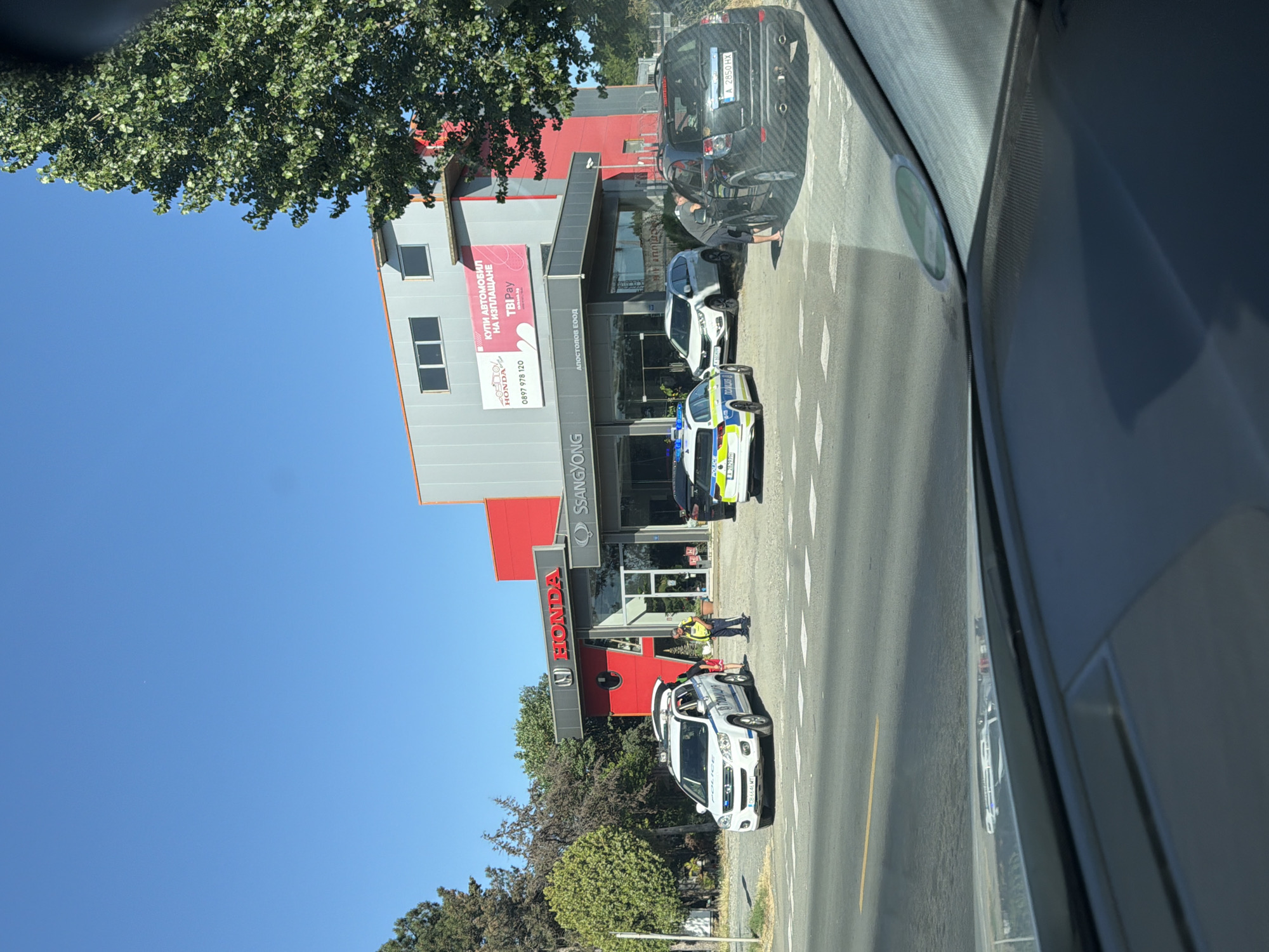 The image captures a scene outside a car dealership in Burgas, Bulgaria. A white police car with illuminated lights and the word "POLICE" in English and Bulgarian text is parked near the entrance. A police officer in uniform, wearing a high-visibility vest, stands beside the open trunk of the police car, interacting with another individual. A white Honda SUV is parked behind the police car. Further to the right, a black SUV is parked in front of the dealership, with a person leaning into its open driver-side window. The dealership building has prominent "HONDA" and "SSANGYONG" signage. The sky is clear and blue, and trees are visible on the left and right sides of the image. The scene appears to be taking place during daylight hours on a street with road markings.