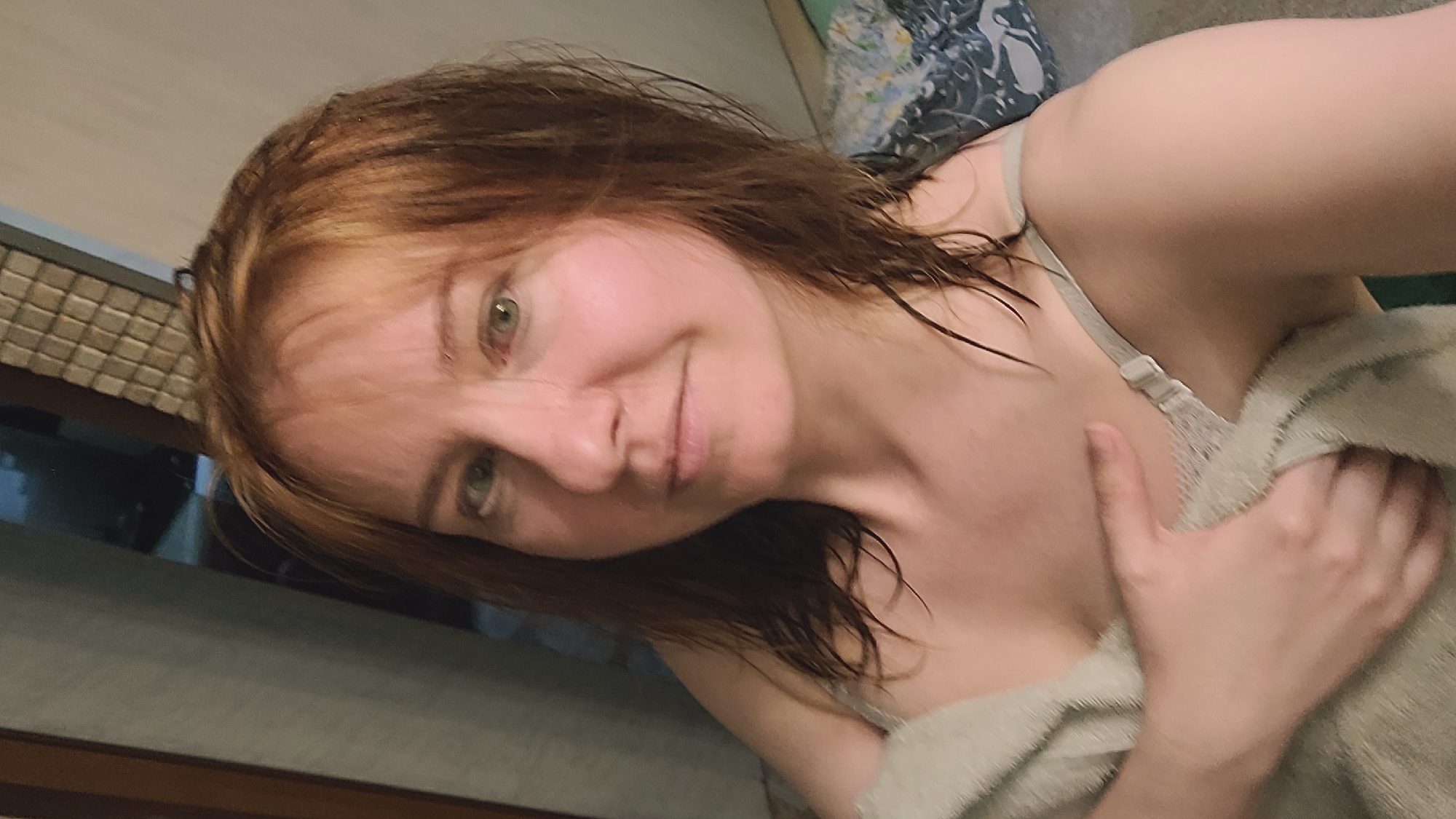 The media file is a selfie of a person with auburn hair and green eyes. The person is wearing a light-colored bra and is wrapped in a light-colored towel. Their hair appears wet, suggesting they may have recently showered or bathed. The lighting is dim, creating a soft glow on their skin and highlighting a slight smile on their face, conveying a relaxed or content emotion. The background is out of focus, but it appears to be an indoor setting, possibly a bathroom or bedroom, with some textured surfaces and a patterned fabric visible. There is no visible text in the media file.