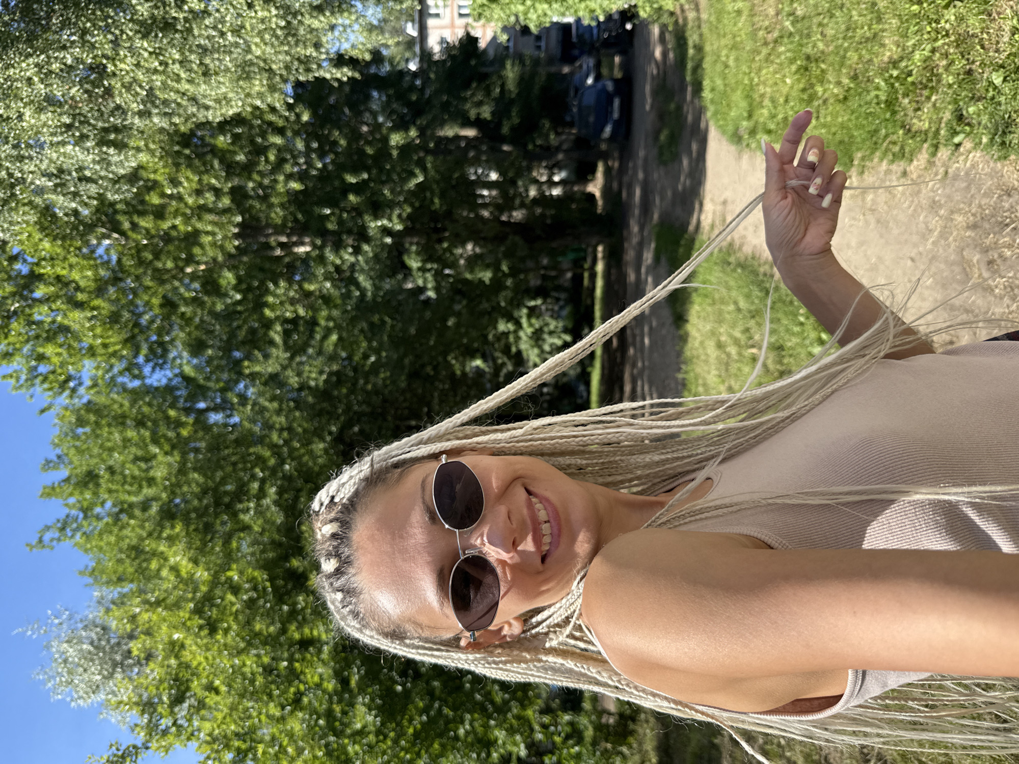 A woman with blonde braided hair and sunglasses smiles at the camera while standing outdoors. She is wearing a light-colored tank top. In the background, there are trees, a path, green grass, and a building with cars parked in front of it. The image appears to be taken on a sunny day. The location is identified as Rusanovo, Russia.