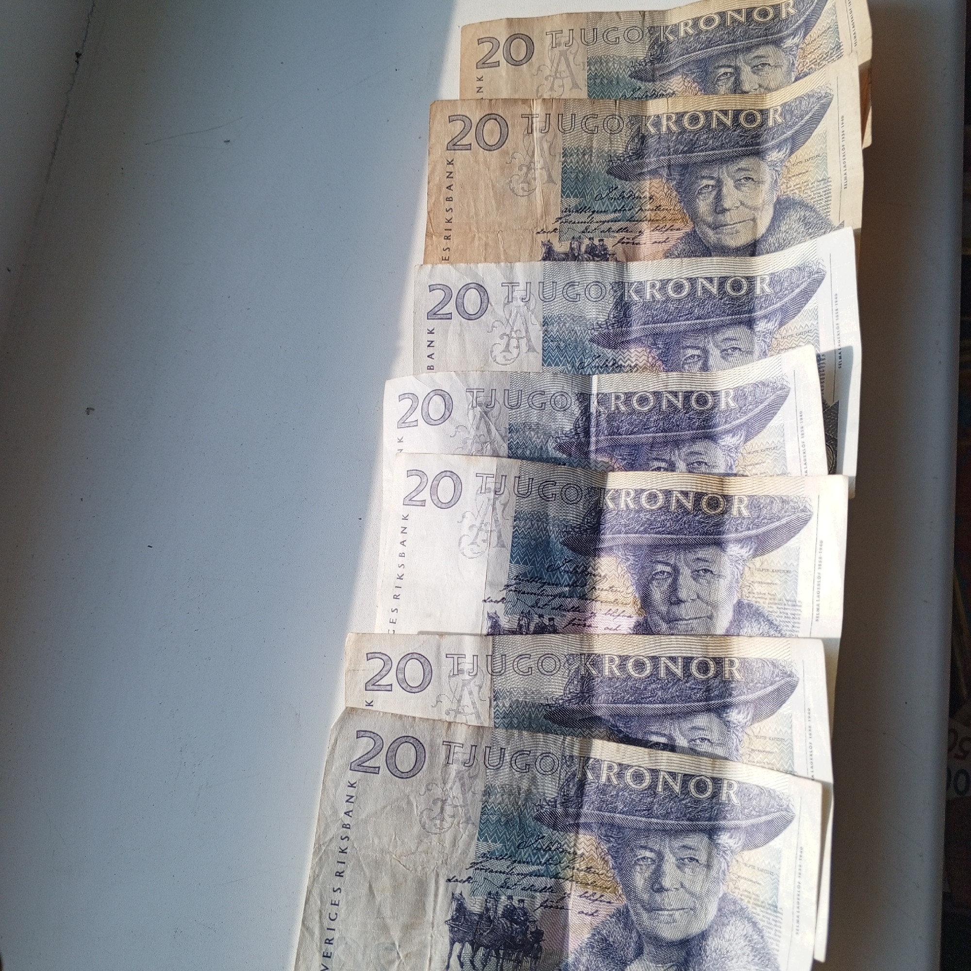 A stack of Swedish 20 kronor banknotes is arranged vertically on a light-colored surface, possibly a table, under natural lighting that casts shadows. The banknotes are fanned out, with the top banknotes partially obscuring the ones beneath. The front of the banknotes features a portrait of Selma Lagerlöf, a Swedish writer, and the denomination "20 TJUGO KRONOR". The background of the banknotes has intricate patterns and text. The surface appears to be a simple, indoor setting. There are no people visible in the image. The image does not contain any information that would allow for the determination of its location in Sayanogorsk, Russia.