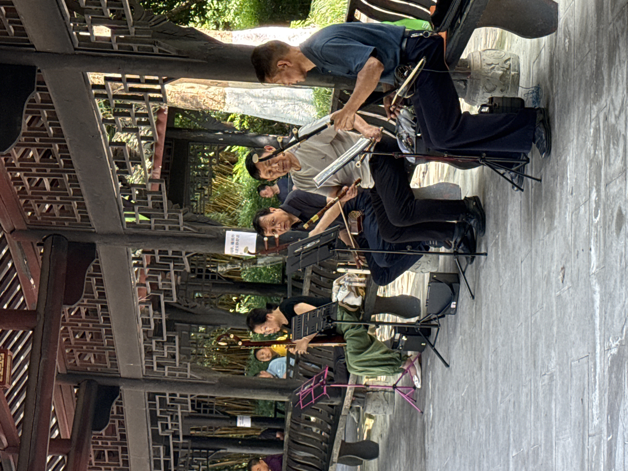 This image depicts an outdoor musical performance by a group of five musicians, primarily men and one woman, in Banan, China. They are seated under a traditional Chinese pavilion, playing what appear to be classical Chinese string instruments.
The musicians are dressed casually; the men wear t-shirts and trousers, while the woman is in a black top and green pants. Several men are playing erhus, identifiable by their bowed strings and distinctive shapes. The woman is playing a plucked string instrument, possibly a pipa or ruan. Each musician has a music stand with sheet music positioned in front of them, suggesting a coordinated performance or practice. Small black speakers or amplifiers are visible on the ground near the performers.
The setting is an intricately designed wooden pavilion, characteristic of traditional Chinese architecture, featuring elaborate lattice work and carved beams on the ceiling and structural supports. The ground is paved with grey stone or concrete slabs. Beyond the pavilion, lush green foliage and trees are visible, indicating a park or garden environment. The scene is well-lit by natural light, suggesting it is daytime with pleasant weather.
A white sign with black Chinese characters is partially visible in the background, containing rules or notices for visitors. The text includes phrases such as "Civilized tourism," "strictly forbidden to make noise," "litter," "do not smoke," and "do not climb," promoting respectful behavior in the public space. The overall impression is one of cultural performance set in a serene, architecturally rich environment.