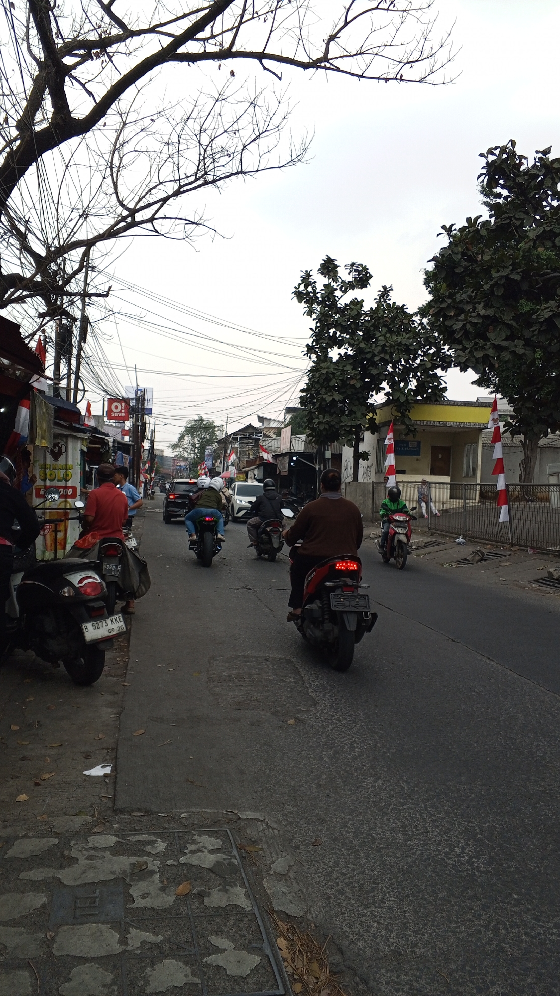 A daytime, eye-level shot captures a street scene in Bekasi, Indonesia, dominated by a two-lane asphalt road. The road is moderately busy with several motorcycles and a few cars. On the left side of the road, there are small shops and stalls, adorned with red and white Indonesian flags. A large tree with bare branches extends from the upper left of the frame, its silhouette stark against the overcast sky, while lush green trees with full foliage stand on the right. Power lines crisscross the sky. Several people are visible, most of them riding motorcycles. One individual in the foreground is on a red motorcycle, facing away from the camera. Further down the road, more motorcycles and cars are in motion. The sidewalks are uneven, with some debris and fallen leaves scattered on the ground. A metal grate is visible on the sidewalk in the lower left corner. The overall impression is a typical urban street in Indonesia, bustling with daily activity.