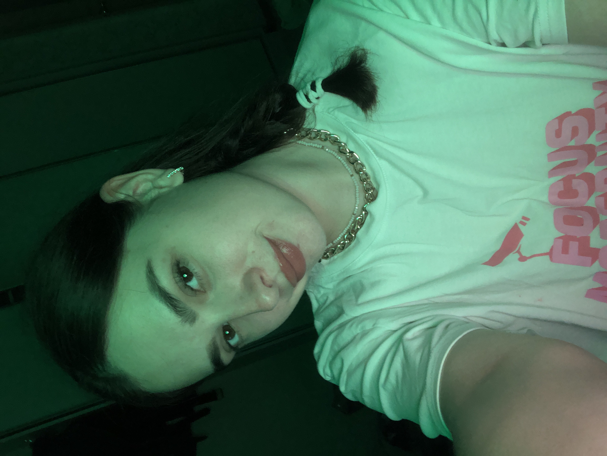In Noviy, Russia, a young woman with dark hair styled in a braid with a black and white tie is captured in a close-up selfie. She is wearing a white t-shirt with a pink graphic that reads "FOCUS" in large letters, partially obscured by her arm. Her face is turned slightly to her left, with her right eye looking directly at the camera. She has on subtle makeup, including lipstick, and wears a thin, sparkly earring in her left ear. Around her neck, she sports two layered necklaces: a thin beaded one and a thicker gold chain. The scene is illuminated by a green light, casting a greenish hue over the entire image and creating a dark, dimly lit environment, typical of an indoor setting. The background is dark and indistinct, offering no specific environmental context within Noviy, Russia.