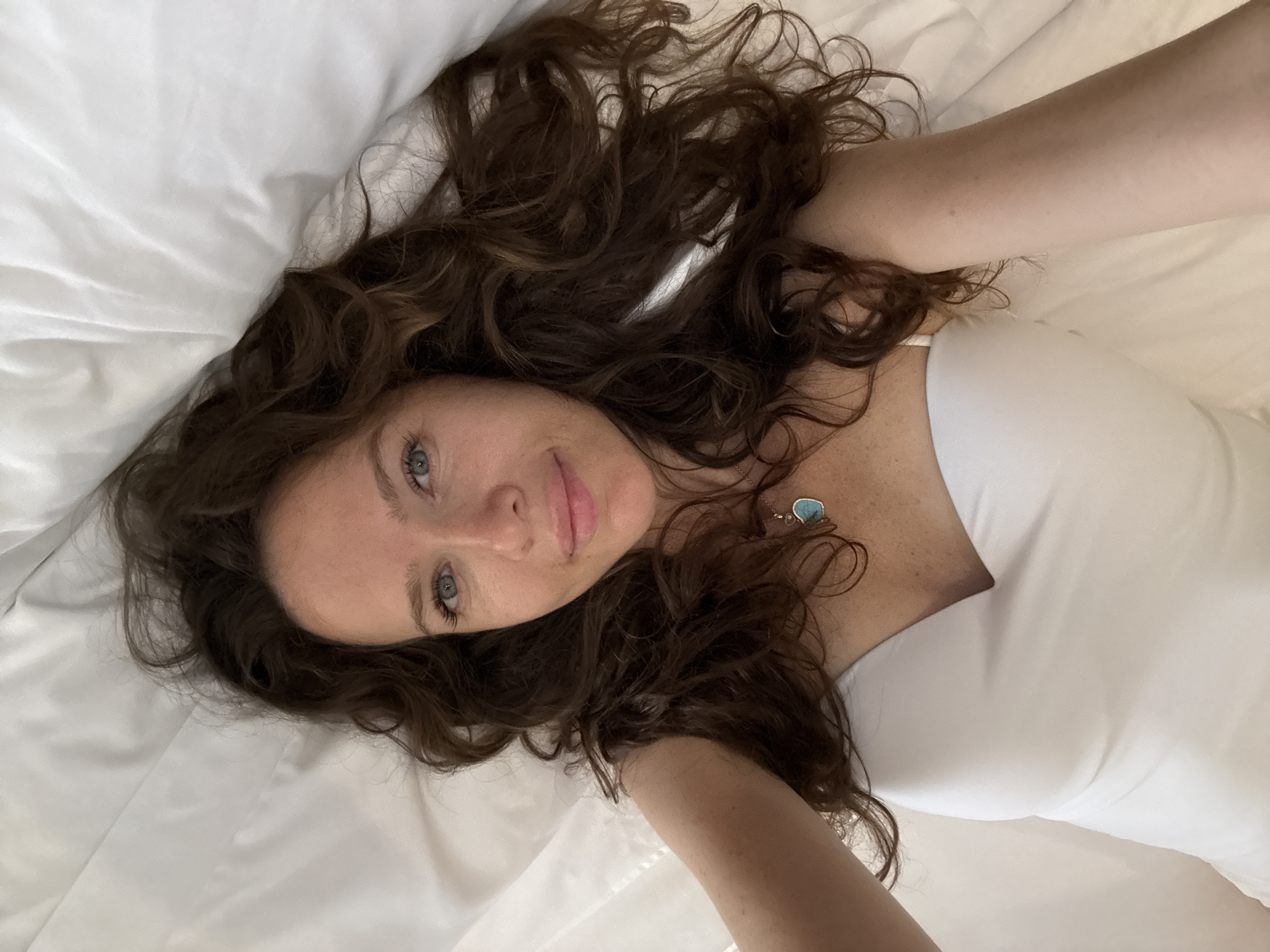 A woman with long, curly brown hair lies on a white, creased surface, likely a bed, and looks at the camera. She is wearing a white strapless top and a necklace with a teardrop-shaped turquoise pendant. Her face is partially illuminated, with her blue eyes and a slight smile visible. The background is a soft, white, textured surface, suggesting an indoor setting. There are no discernible elements in the image that can be used to determine its location within the city of Dubai, United Arab Emirates.