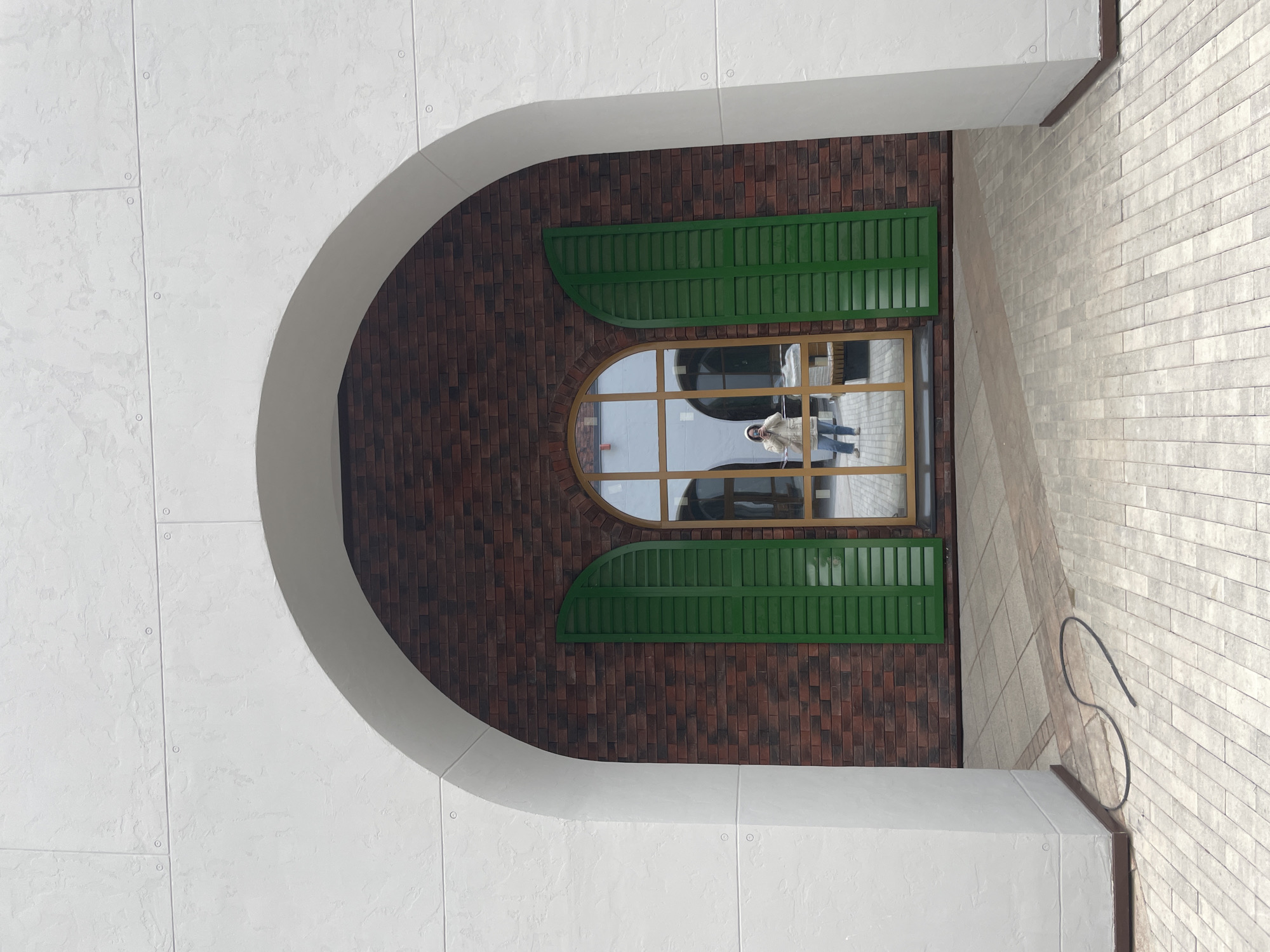 The image captures a detailed view of an arched architectural feature, likely an entrance or a passage. The outer structure on the left consists of a white, textured facade made of large panels with visible circular fasteners. This white wall forms a prominent archway.
Through this archway, a reddish-brown brick wall is visible, serving as the backdrop for a large window. The window has a distinctive curved top, complementing the arch above it, and features a golden-brown frame divided into multiple panes. Flanking the window are two vibrant green, louvered shutters, which also have curved edges to integrate smoothly with the overall design.
Reflected clearly in the window is a person, likely the photographer, wearing a light-colored jacket or coat, blue jeans, and a white head covering (possibly a hat or hood). The reflection also shows parts of another building or structure with large, circular window-like elements.
To the right of the main arched opening, a wall constructed from light-colored, rectangular tiles or bricks extends into the scene. The ground in this area is also paved with similar light-colored tiles. A coiled black cable or hose is visible on the tiled ground near the right wall.
The scene is illuminated by bright daylight, suggesting good weather conditions. There is no visible text in the image. The architectural style blends modern elements (white panels, sleek curves) with traditional materials (brick, louvered shutters). The location context is Izhevsk, Russia.