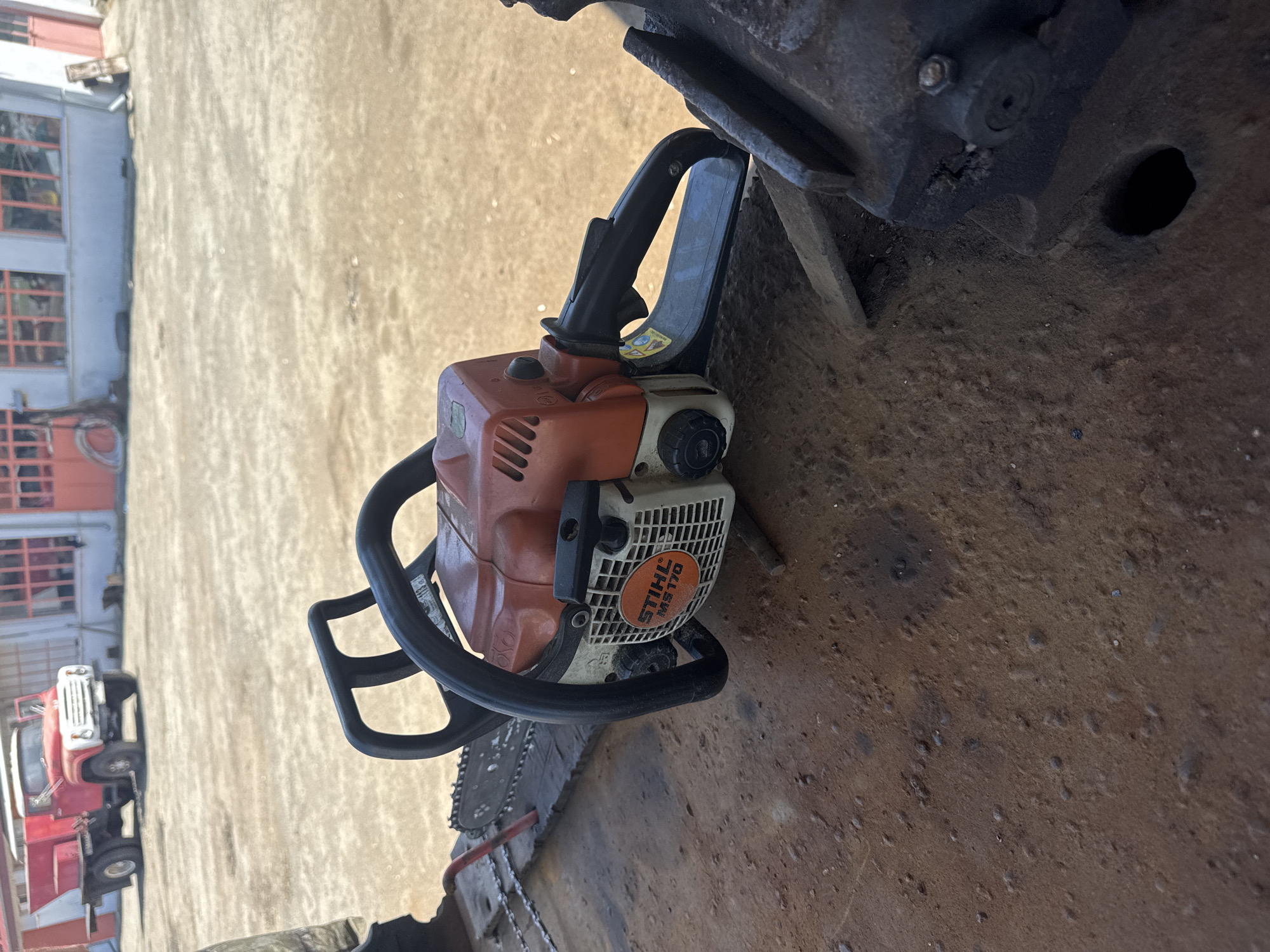 The image depicts a Stihl MS 170 chainsaw lying on a gritty, possibly concrete surface, partially under a dark, metal object with a circular hole. In the background, a light-colored building with orange-framed windows and a red truck are visible. The location is Sarnevo, Bulgaria. There are no people or discernible interactions visible in the image.