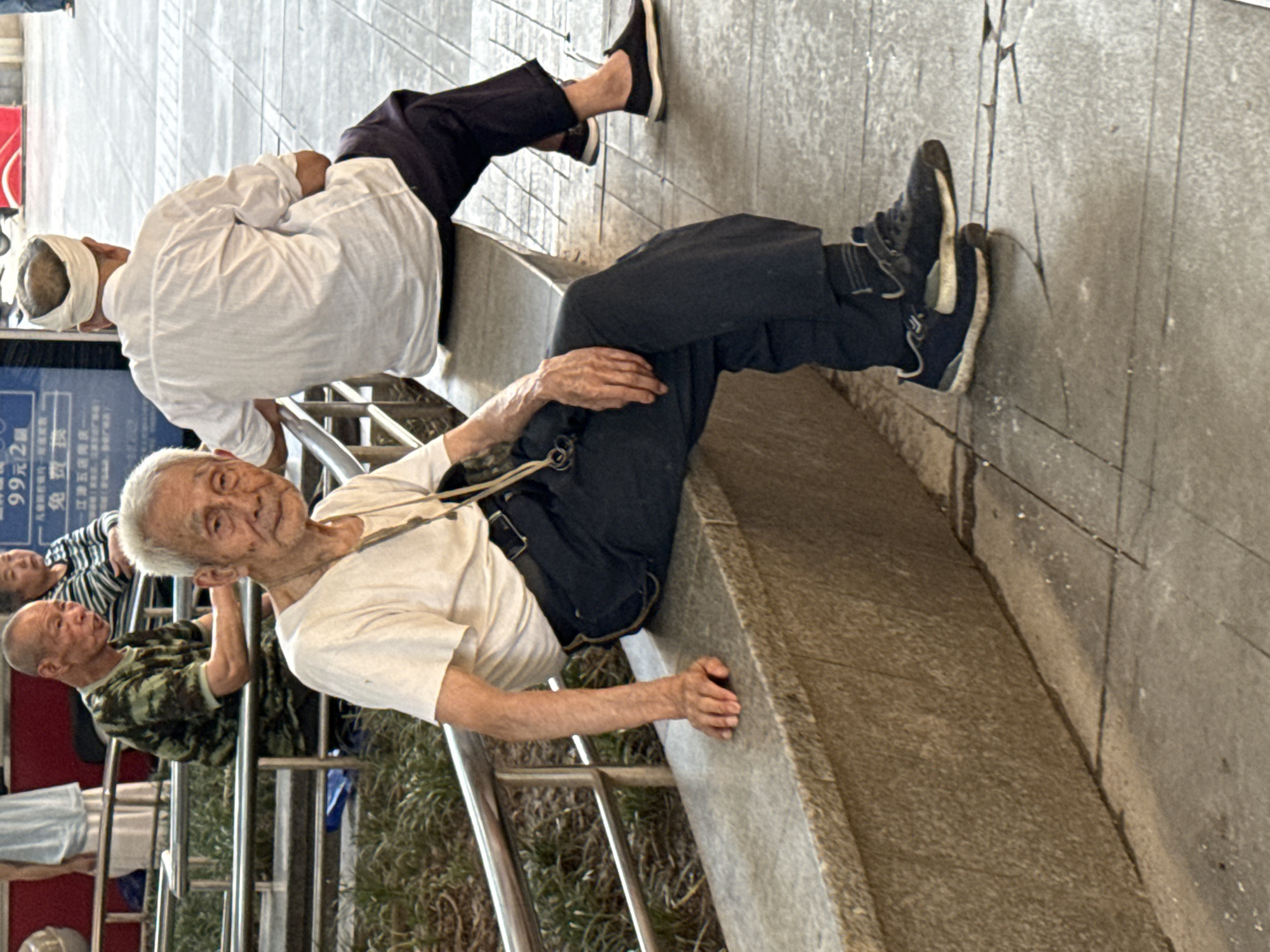 This image, taken in Jiangjin, China, depicts an outdoor public space populated by several elderly individuals during what appears to be a bright day.
In the foreground, an elderly man with white hair sits casually on a low stone ledge. He wears a white short-sleeved shirt, dark trousers, and dark sneakers, looking directly at the camera with a friendly, subtle smile. His hands, showing prominent veins, rest on his lap and the ledge. A dark pouch or bag hangs from a strap across his chest.
Behind him and slightly to his left, another elderly man in a white long-sleeved shirt and a white head wrap is seated on a higher section of the same stone structure, facing away from the camera. Further left, an older man with a shaved head and a camouflage t-shirt leans on a metal railing, looking towards the foreground. Partially visible behind the railing are other people, including one in a striped shirt and another in a light blue top.
The setting features tiered stone or concrete structures, likely steps or seating areas, along with metal railings that separate different sections of the space. Green foliage is visible beyond the railings. The ground is paved with a light-colored stone, exhibiting some cracks. In the background, a blue sign displays Chinese characters, including "99元2" and "免费" (meaning "free"), providing a local contextual detail. The scene captures a moment of calm, with people resting and observing their surroundings in a communal outdoor area.