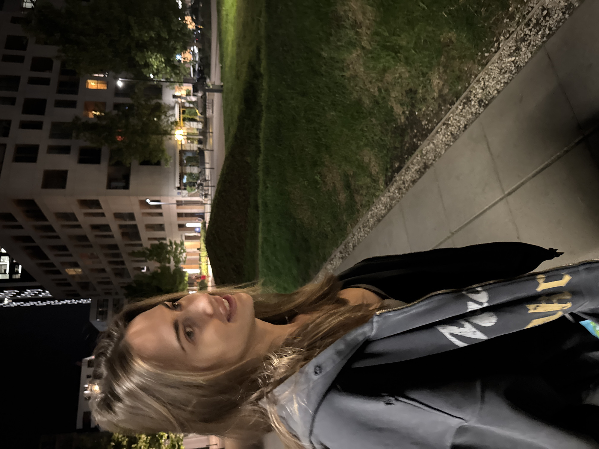 A woman is standing on a sidewalk in an urban environment. To her left is a building with multiple windows, illuminated from within. Adjacent to the sidewalk is a grassy area. The scene appears to be captured at night.
