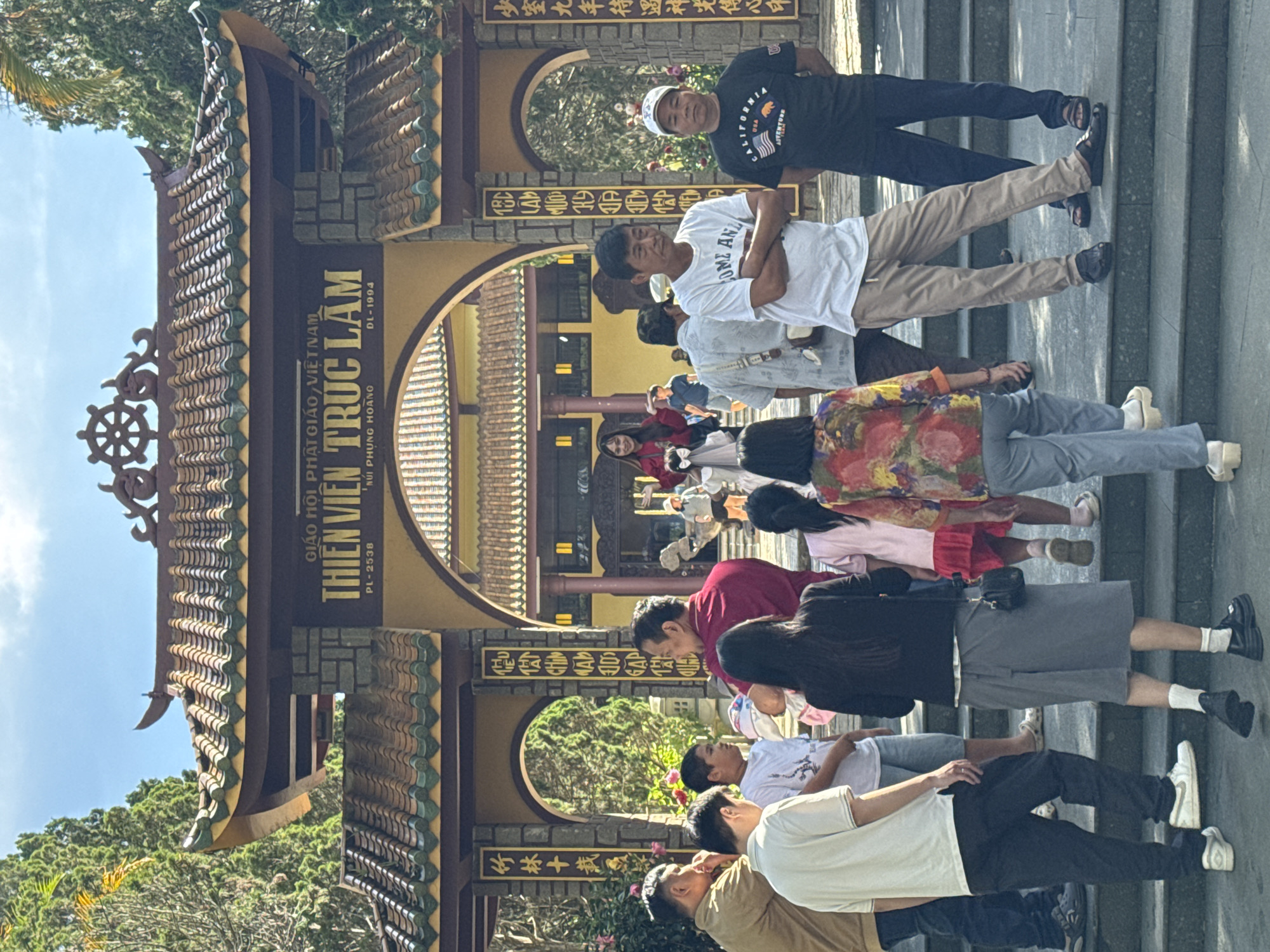 The image shows the entrance to Thien Vien Truc Lam, a Buddhist monastery in Da Lat, Vietnam. A group of people, dressed in casual clothing, are gathered on the steps leading up to the entrance. The architecture of the entrance is traditional Vietnamese, with a tiled roof and ornate carvings. The monastery appears to be situated in a lush, green environment, with trees and foliage visible in the background. The sky is a clear blue, suggesting a sunny day. The people are standing and walking, some are looking towards the entrance, while others are engaged in conversation. The overall scene depicts a tourist attraction or a place of worship that is open to visitors.