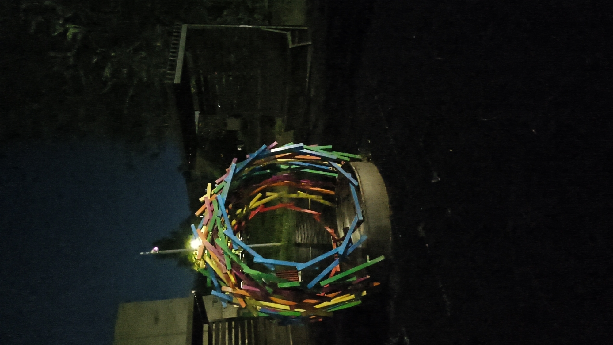 In the city of Mikhaylovka, Russia, an art installation resembling a colorful, spherical nest is displayed outdoors at night. The structure is composed of numerous brightly colored wooden sticks, primarily in shades of green, blue, yellow, and red, arranged to form an open, hollow sphere. The installation rests on a circular, metallic base. In the background, a dark, leafy tree looms, and to the left, a portion of a building with a slanted roof is visible. A flag pole with a small flag, illuminated from below, stands near the building. The ground surrounding the installation is dark and appears to be unpaved. To the right of the art piece, a dark, enclosed structure with a slatted roof and a single bench is visible. The scene is illuminated by ambient light sources, suggesting it is a public space, possibly a park or an exhibition area. No individuals are present in the image.