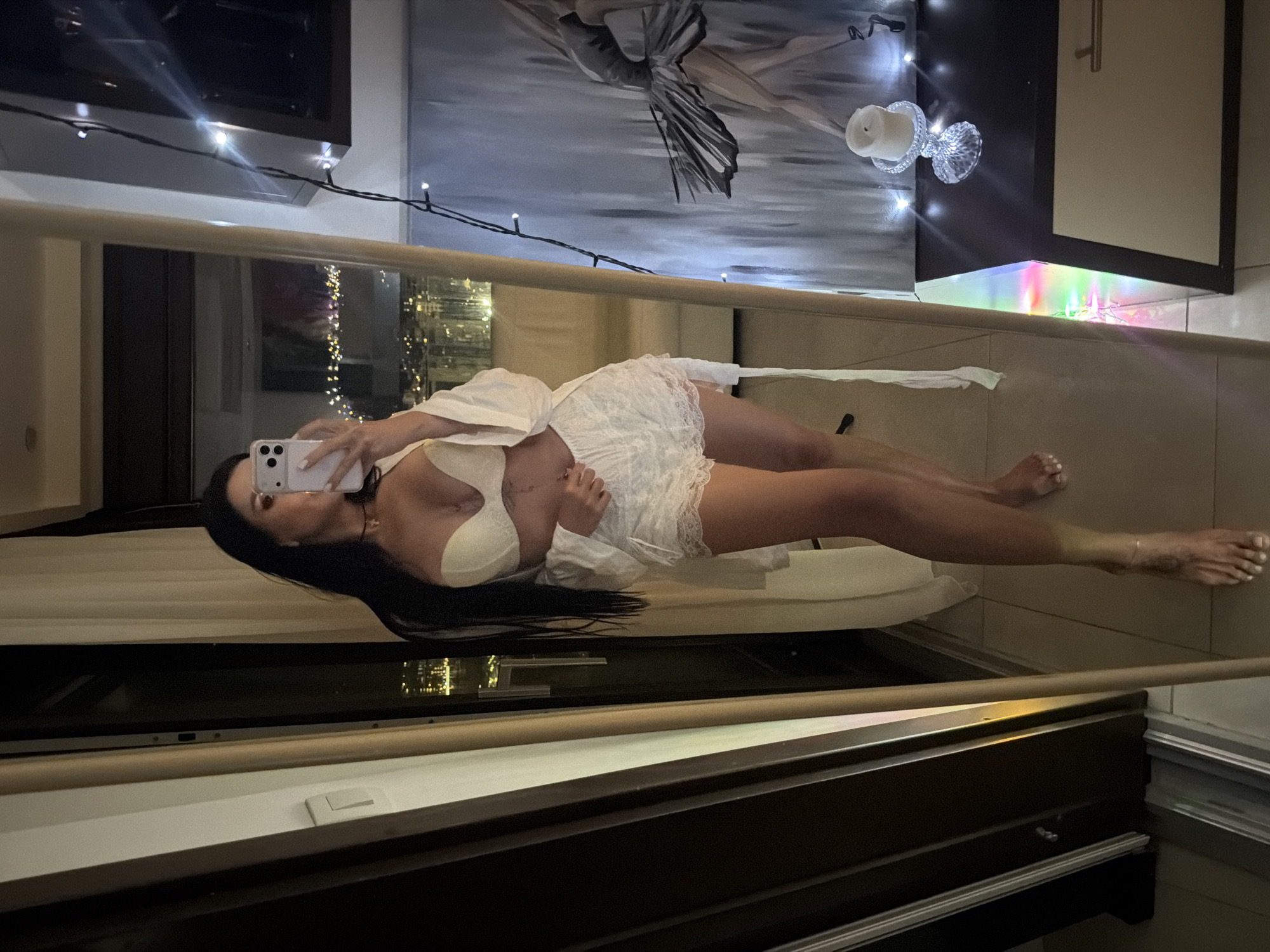 A young woman with long, dark hair is depicted taking a mirror selfie in what appears to be an apartment or hotel room in the city of Boreti, Montenegro. She is standing barefoot on a light-tiled floor, wearing a light-colored strapless bra, white lace shorts, and an open, long-sleeved white robe. A small chest piercing is visible, along with a dark choker necklace featuring a small cross pendant. She holds a light-colored smartphone with both hands, pointing it towards her reflection in a large, full-length mirror that dominates the right side of the frame. Behind her, a large glass door or window overlooks a city at night, with numerous illuminated buildings visible in the distance. To the immediate left of the woman, light-colored curtains are partially drawn. To the right of the mirror, a dark console table or dresser holds a white candle in an ornate clear glass holder, surrounded by a string of small white LED lights. Behind the table, a framed artwork featuring a figure, possibly a dancer, in muted tones is partially visible. Another string of multicolored LED lights is placed along the base of the console table.