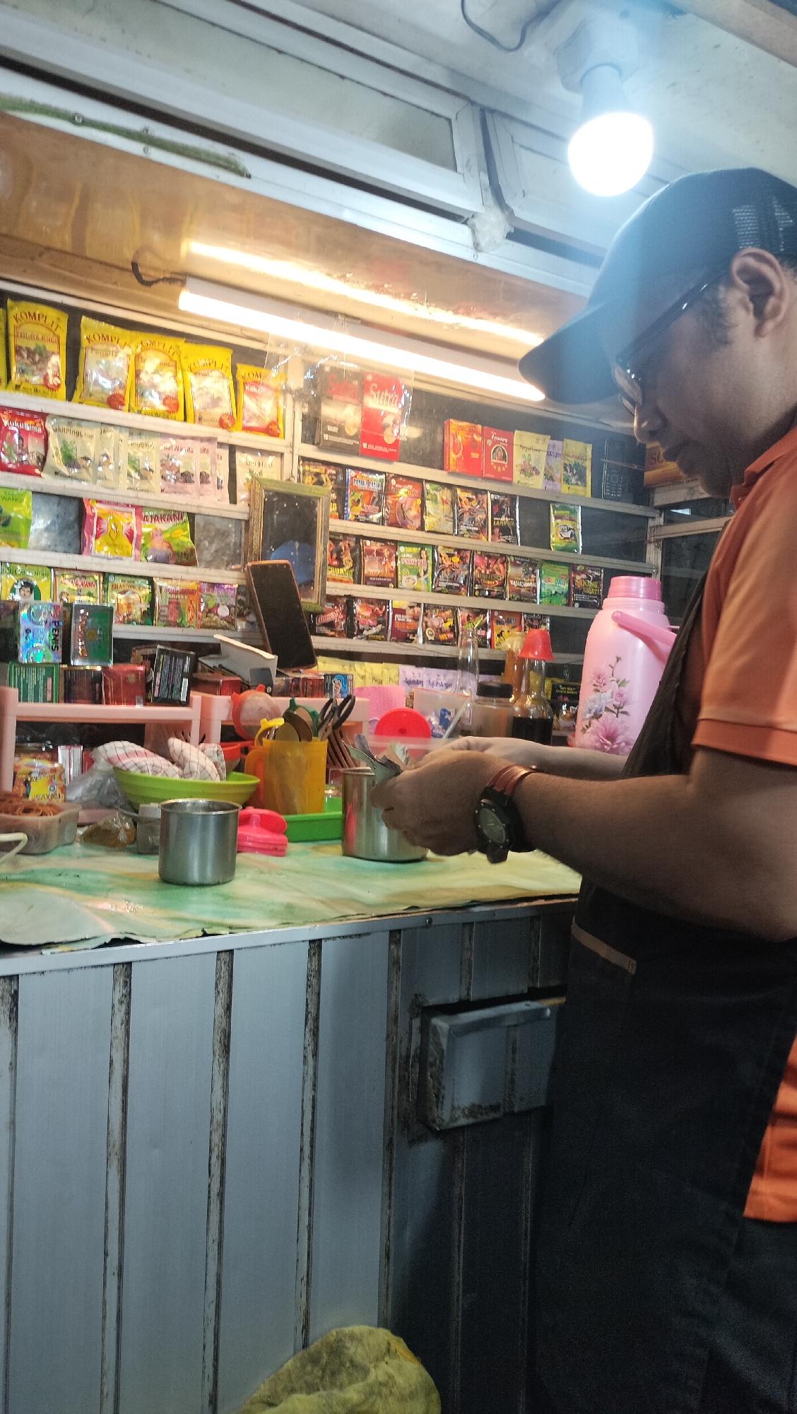 The image depicts a man wearing an orange shirt and a black baseball cap, standing behind a counter in what appears to be a small shop or street vendor stall in Lhokseumawe, Indonesia. The stall is filled with rows of packaged goods, many of which are colorful food items or snacks. The man is engaged in an activity that suggests he is handling money or preparing a transaction, as his hands are close to a stainless steel container and he appears to be holding something within them. A bright light bulb illuminates the scene from above. Various items are visible on the counter, including bowls, utensils, and bottles, contributing to the impression of a functional retail space. The background shows a densely packed shelf of products, typical of a small convenience store or a roadside stall catering to local consumers.