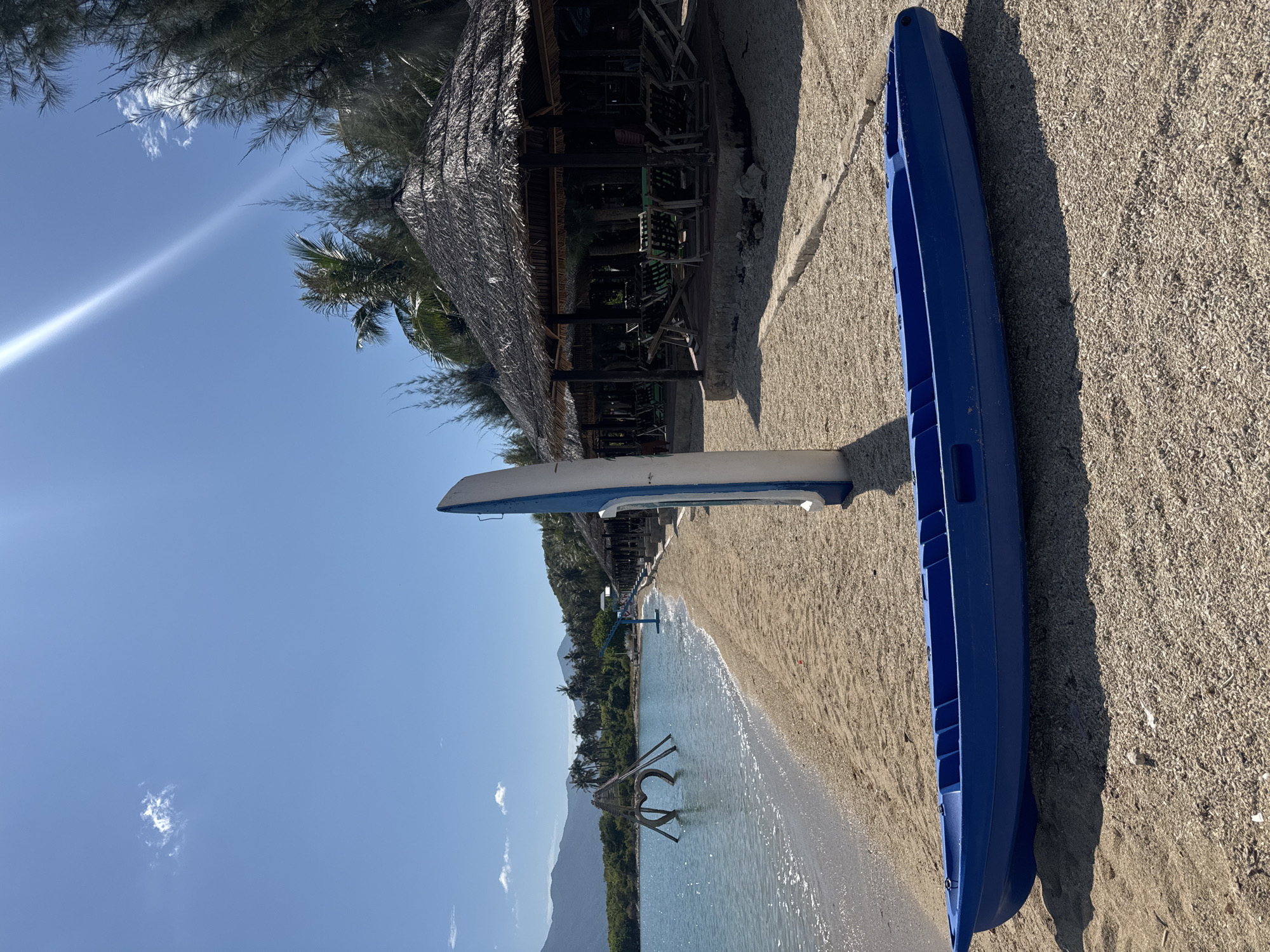 A blue kayak rests on a sandy beach in Ninh Ích, Vietnam. To the left, the clear turquoise ocean meets the shore, with a wooden sculpture resembling a heart visible in the water. Palm trees and other lush vegetation line the background, bordering a row of thatched-roof huts with lounge chairs. The sky above is a bright, clear blue with a single, wispy cloud. The sun casts a strong glare and shadows across the scene.