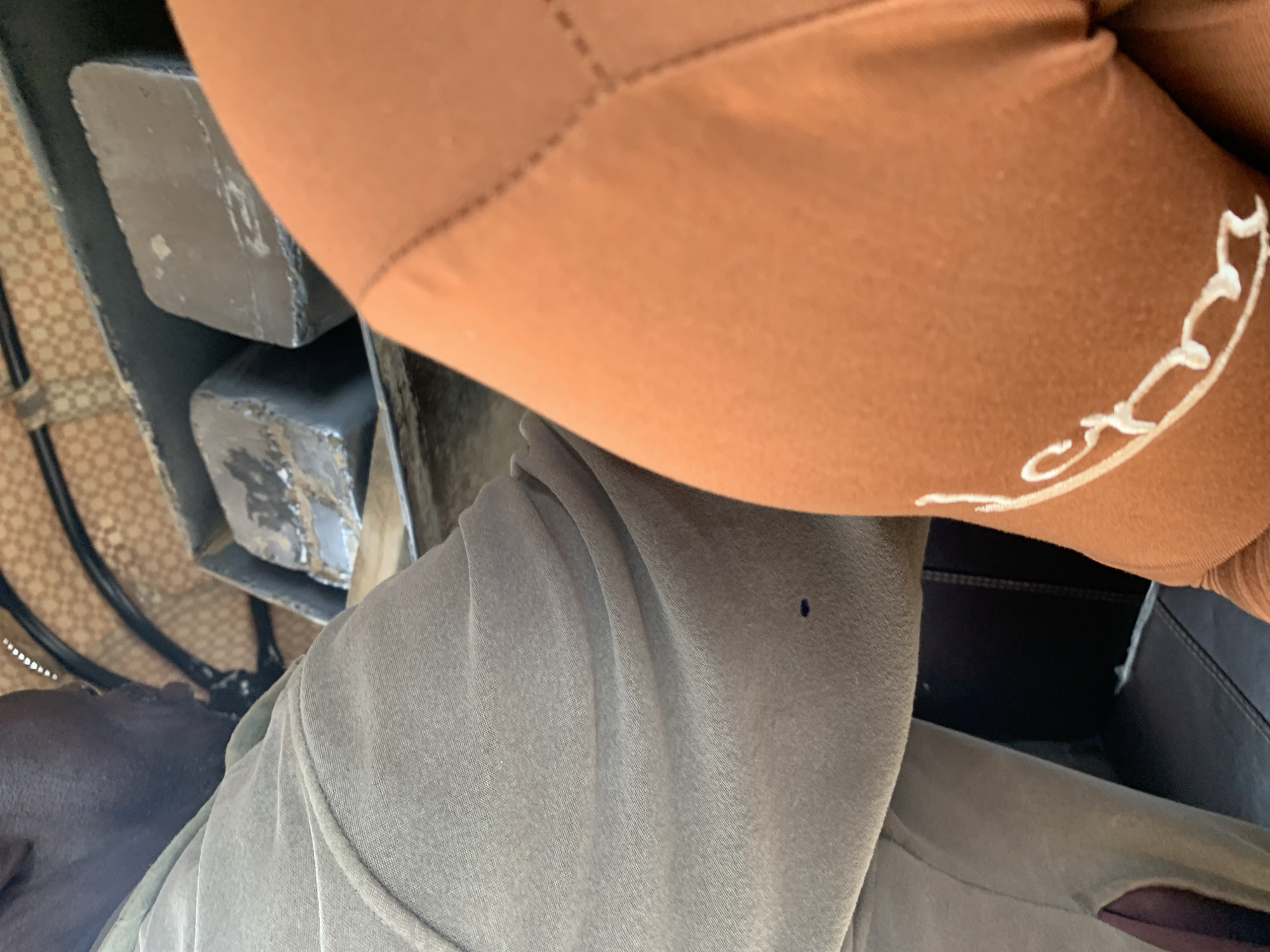 The image presents a close-up interior scene, likely within a vehicle, situated in the city of Rafin Par, Nigeria. On the left, a person's upper back, side profile of the head, and left arm are visible. The individual has dark skin and short dark hair. Their left arm and shoulder are covered by a light grey long-sleeved garment made of a soft, possibly fleece-like fabric. A small, dark blue dot is present on the fabric of the left arm, approximately mid-forearm.
On the right side of the frame, the shoulder and upper arm of a second person are visible, clad in an orange-brown garment. The fabric features distinct stitched seams running diagonally across the upper shoulder area. White, stylized, curvilinear embroidery is present on the lower portion of this garment, curving upwards from the bottom right.
Between the two individuals, a dark, recessed platform or storage area is visible. Within this area, two dark grey, rectangular, solid blocks are placed side-by-side. These blocks exhibit some surface imperfections and reflections from an overhead light source. Behind these blocks, a light tan or beige patterned surface is visible, possibly part of the vehicle's interior trim or ceiling, displaying a repeating geometric grid pattern. Further down on the left, beneath the left individual's arm, a dark vertical panel and a dark horizontal surface are discernible.