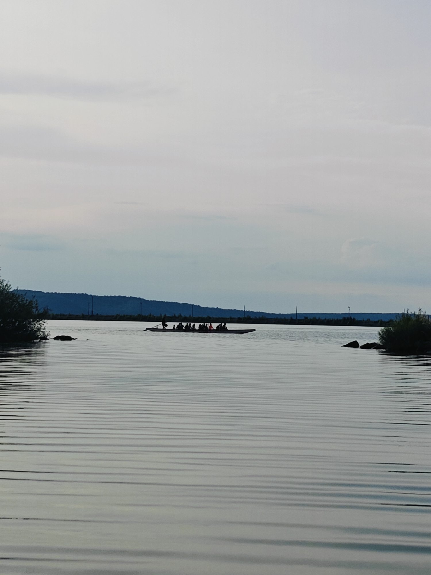 The image captures a wide shot of a body of water, likely a river or a lake, under an overcast sky. In the middle distance, a long, narrow boat is visible, carrying multiple people, appearing to be engaged in rowing or a similar activity. Their silhouettes are dark against the lighter water. The water itself exhibits distinct, horizontal ripples, suggesting a gentle current or breeze. The background features a dark, undulating treeline or hillside, dotted with faint, vertical structures that could be utility poles. On the left and right sides of the frame, the edges of dense foliage are present, framing the central scene. The overall impression is one of a serene outdoor activity taking place in a natural setting. This scene is representative of the Kama River or a reservoir near Chaikovsky, Russia, a city known for its industrial facilities and recreational areas along the Kama River.