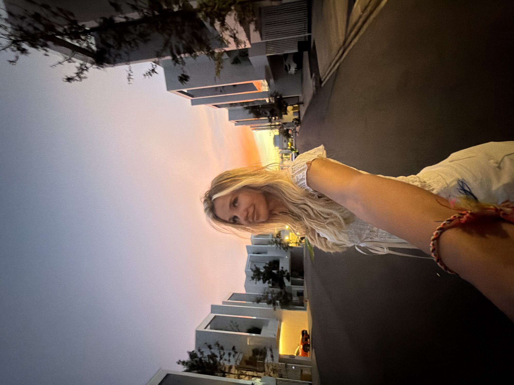 This is a selfie taken by a woman with long blonde hair, wearing a white dress. She is standing on a street with modern architecture, possibly a residential area, with buildings on either side of the road. The sky is illuminated with the colors of a sunset, displaying shades of pink, orange, and yellow. A red car is parked in the driveway of one of the houses. The woman's arm is extended towards the camera, and she is wearing a colorful braided bracelet on her wrist. The scene suggests a warm, late afternoon or early evening setting in an urban environment. The location is identified as the city of Thep Krasatti, Thailand.