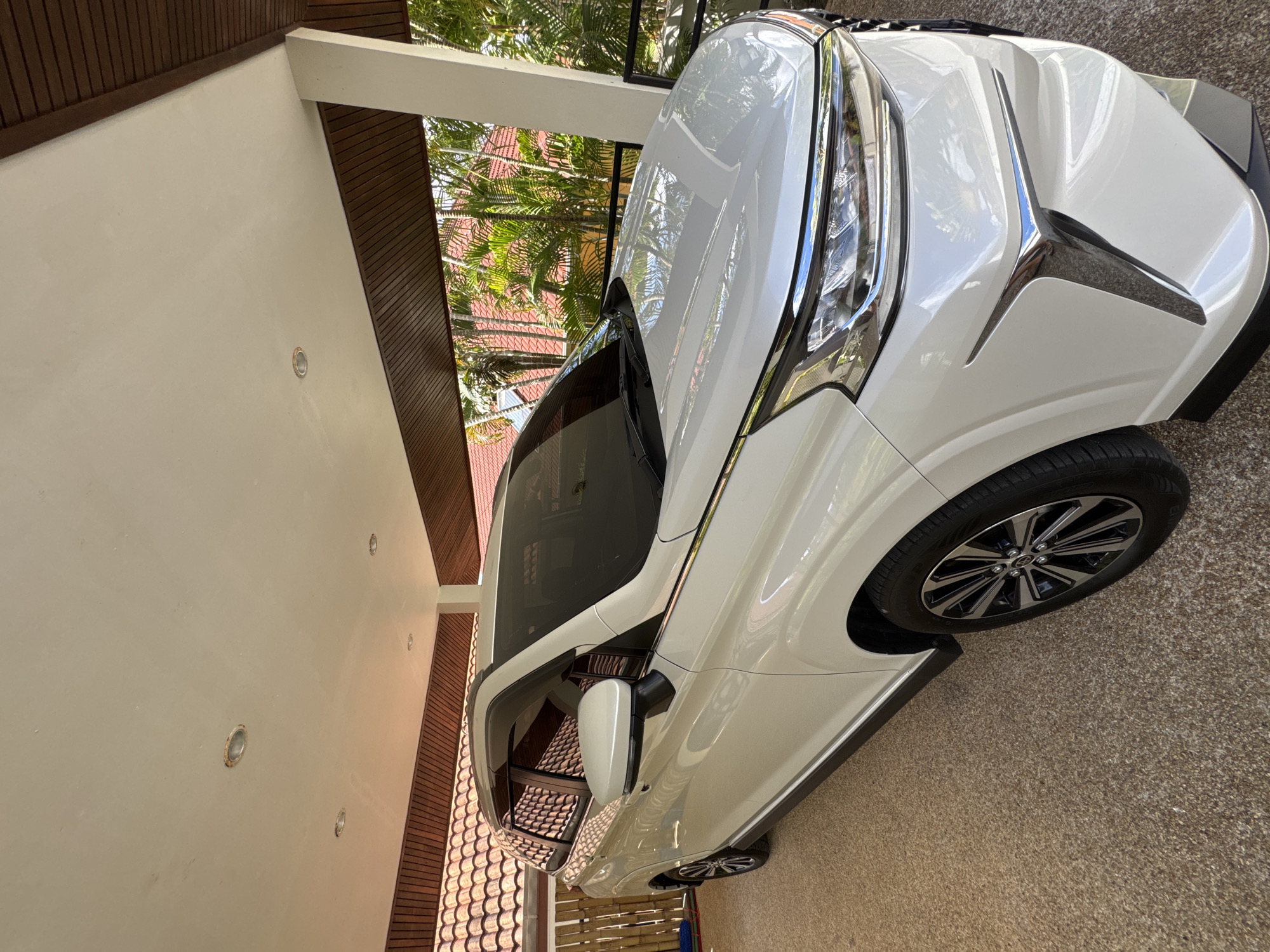 The image depicts a white car parked in what appears to be a driveway or garage. The setting seems to be under the roof of a building with white walls and dark brown wooden trim. The presence of tropical plants and rooftops visible through the building's support beams suggests a warm climate. The car is the main subject, showing its front right side, including the headlight, wheel, and a portion of the hood. The ground surface is a light brown color, possibly concrete or a similar material. There is no visible indication to confirm the specific location of Rawai, Thailand.