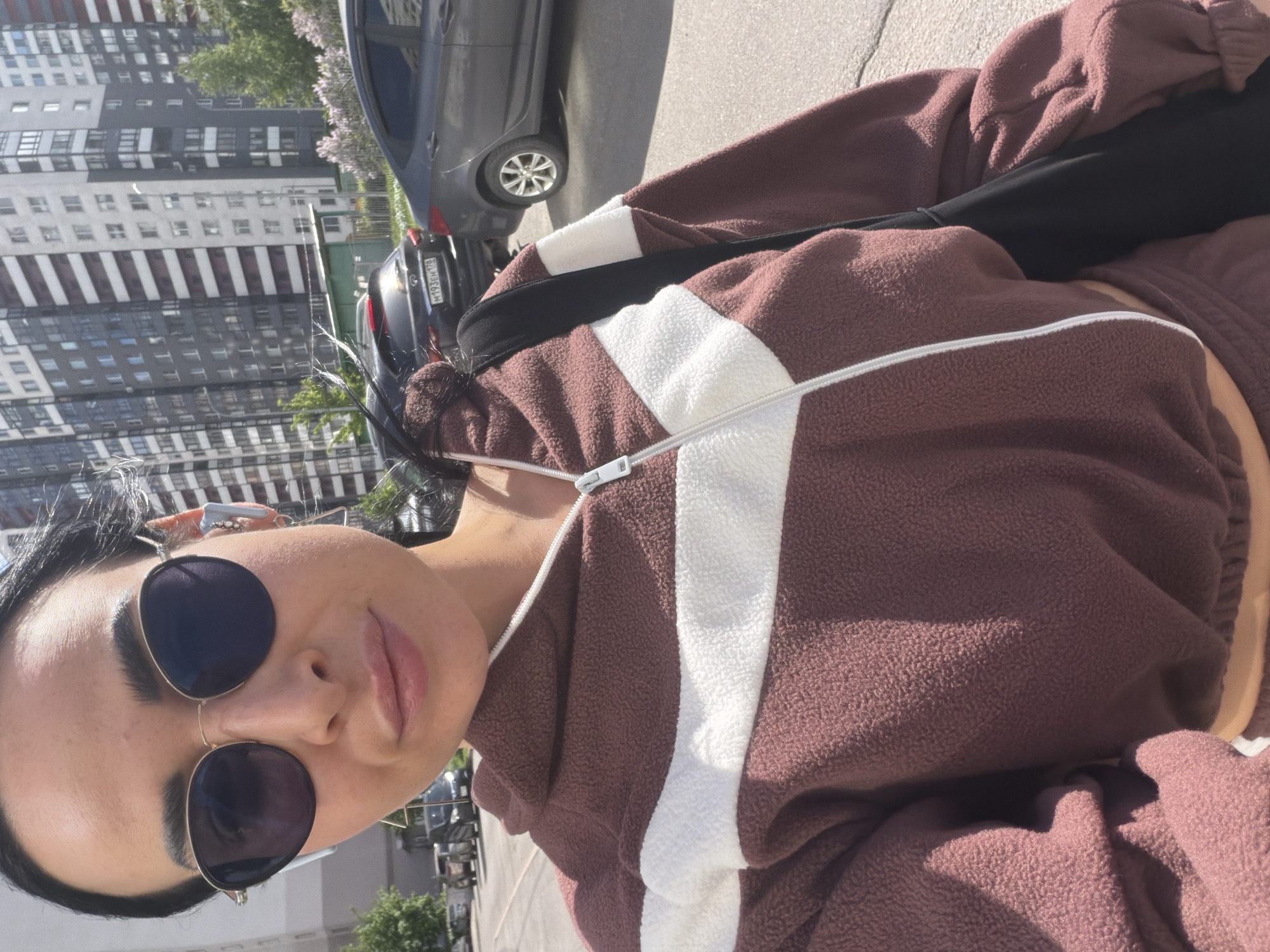 A selfie shows a woman with dark hair and sunglasses, wearing a brown and white jacket with a visible zipper, standing outside in Saint Petersburg, Russia. The background includes a gray car, and in the distance, tall residential buildings.