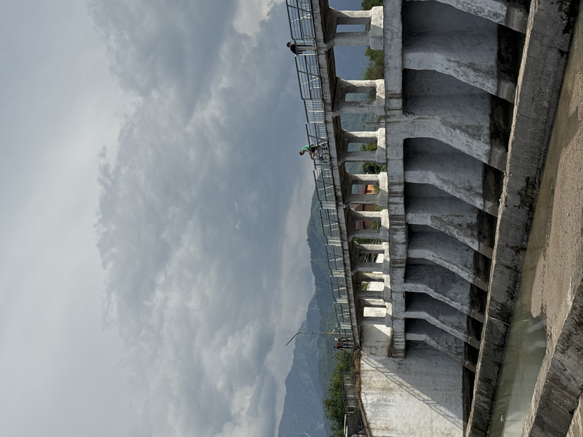 A low-angle, vertical shot captures a section of a concrete dam against a cloudy sky. The dam's structure is characterized by repeating arch-like openings and thick concrete supports. A metal railing runs along the top edge of the dam.
Several people are visible on the dam's walkway. In the middle distance, one person on a bicycle is bent over, appearing to be in motion. Further along the walkway, another person stands near the railing. Towards the foreground, a third person is also near the railing.
Beyond the dam, a lush green mountain range is visible under the overcast sky. The overall scene suggests an outdoor recreational setting, possibly a park or a scenic viewpoint. The time of day appears to be daytime, with diffused light due to the clouds. No visible text is present in the image. The activity depicted is casual, with people enjoying the view or cycling on the dam.