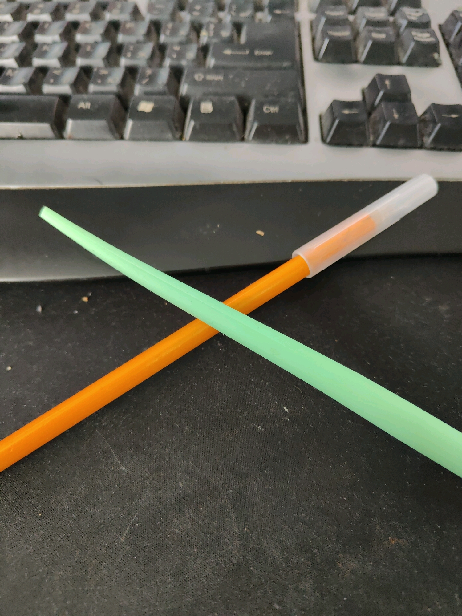 Two brightly colored thin rods, one orange and one light green, are laid diagonally across a dark, textured surface, forming an 'X' shape. The orange rod is positioned in the lower left to upper right quadrant, while the green rod extends from the upper left to lower right. A transparent plastic cap, likely for a pen or marker, is fitted onto the top right end of the orange rod, revealing an orange interior. The background consists of a blurry, grey and black keyboard with visible keys. The scene is indoors, on a desk or similar surface, likely in a home or office environment in Homyel, Belarus.