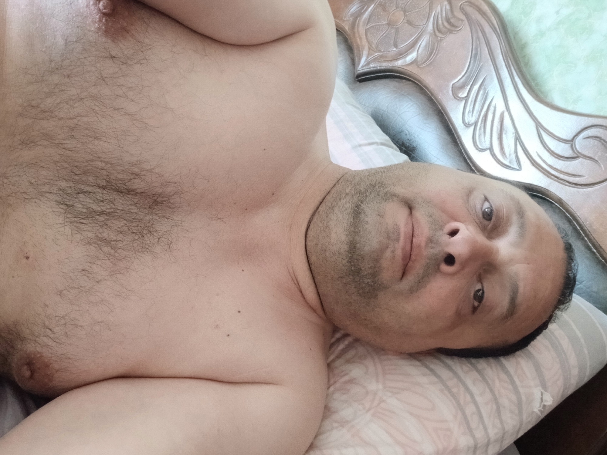 A medium-shot selfie shows a man lying on his back on a bed. He is shirtless, with dark hair on his chest and abdomen. His head is turned towards the camera, and his eyes are open. He has a beard and appears to be in his 40s or 50s. The background consists of a pillow and a headboard. The scene is a bedroom, likely in Telavi, Georgia, judging by the provided context.