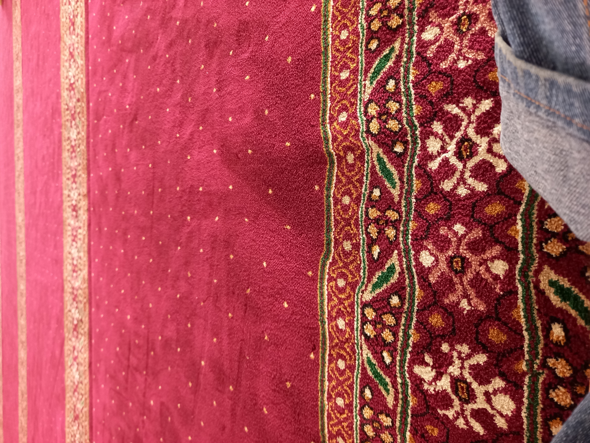 A close-up, low-angle shot shows a maroon carpet with a repeating pattern of gold dots and a decorative border. The border features intricate geometric designs in gold, green, and white. A portion of blue denim jeans is visible in the lower right corner of the frame, suggesting a person is present. The lighting is soft and diffused, creating a warm ambiance. The scene is likely indoors, given the carpet and the close framing. It is not possible to determine the specific location in Samarinda, Indonesia, from the visual information provided.