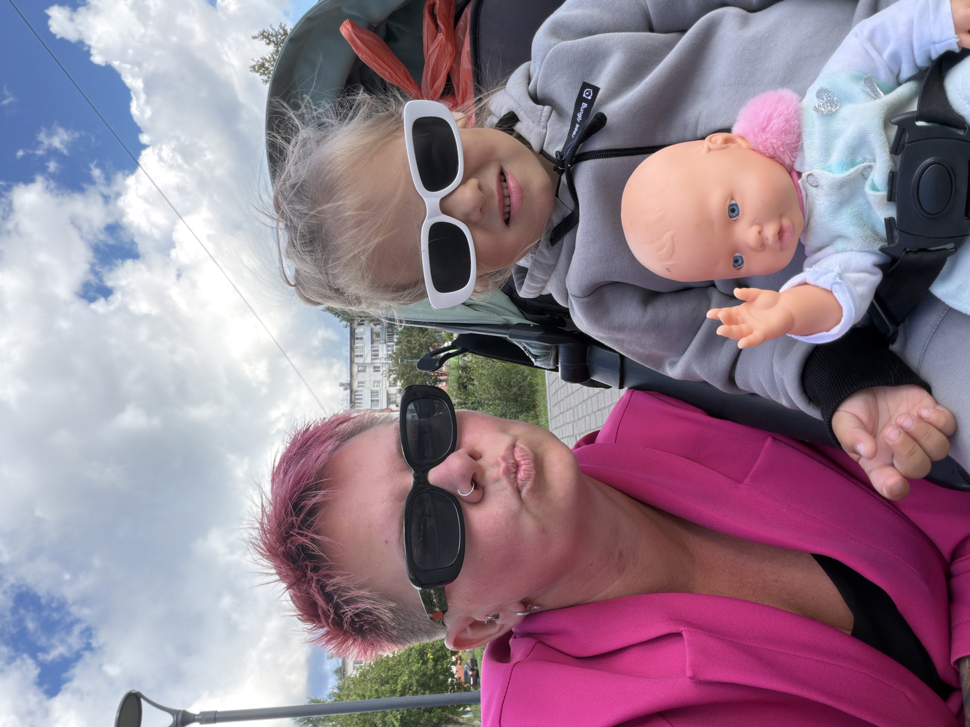 A woman with spiky pink hair and black sunglasses makes a kissing gesture towards the camera, her lips puckered and a silver hoop earring visible. She wears a bright pink blazer over a black top. To her right, a young girl with blonde hair also wears sunglasses, these white-rimmed, and a grey hoodie. The girl holds a baby doll with blue eyes and a pink fuzzy collar, its right arm raised in a wave. They are seated in a stroller, the seat appearing to be a neutral grey. In the background, buildings and trees are visible under a cloudy sky. The scene appears to be outdoors in a public area, possibly a park or street, in the city of Chaikovsky, Russia. A lamppost is visible to the far left.