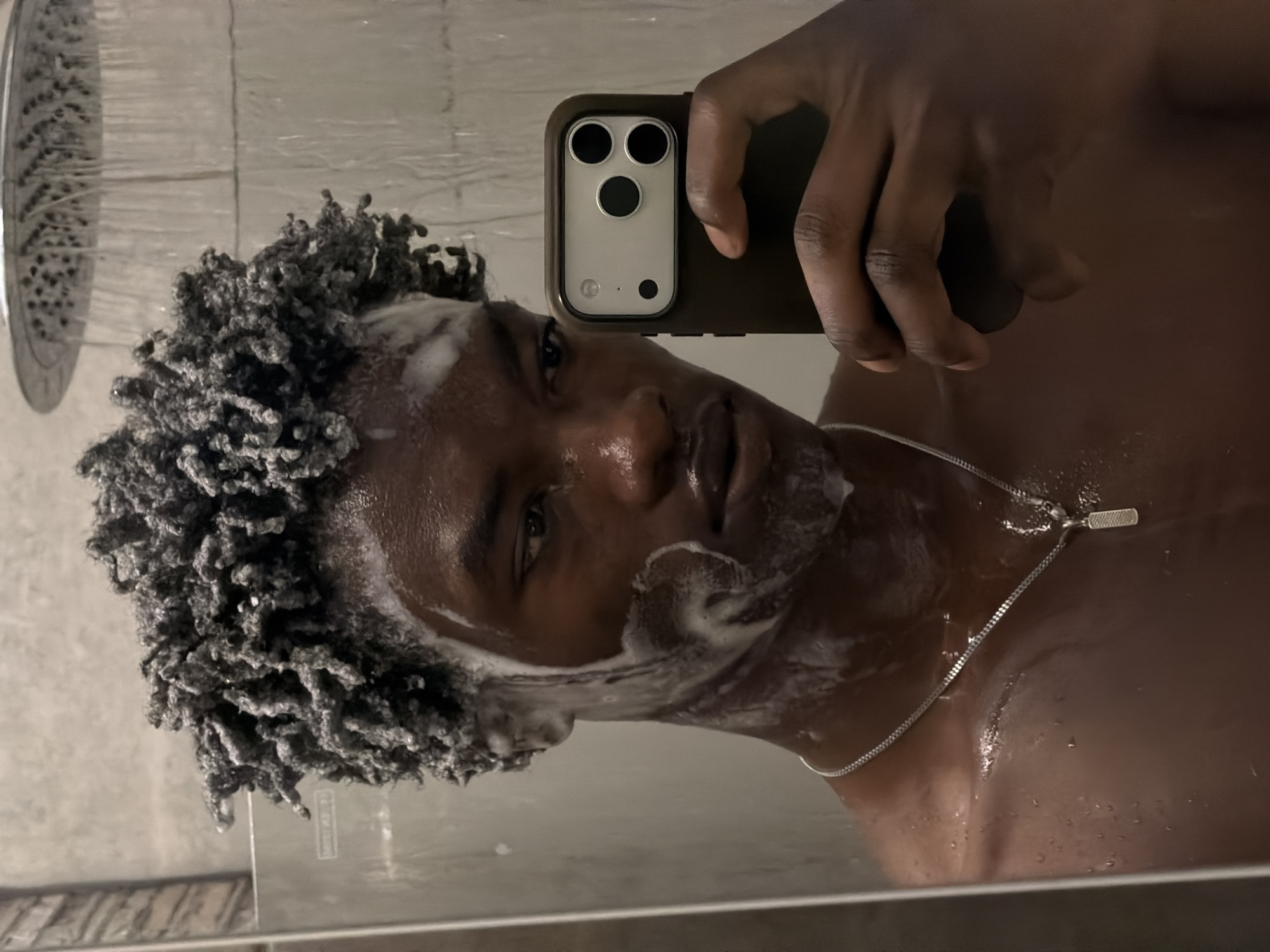 A young adult male is captured in a selfie taken through a reflective surface, likely a mirror or glass shower door, while actively showering. His dark, textured hair and parts of his face are covered in white suds and foam, indicating he is washing. He wears a thin silver chain necklace with a small, elongated rectangular pendant. His right hand holds a smartphone, visible with a dark case and a three-lens camera module with a flash, characteristic of a modern iPhone model. In the upper left background, a showerhead is actively spraying water. The setting is an indoor bathroom, specifically within a shower enclosure, with light-colored walls or tiles visible through the water and steam. The individual's expression is neutral as he looks towards the camera. The lighting suggests an indoor artificial light source. No specific time of day or external location cues are discernible from the image content. No visible text appears in the image.