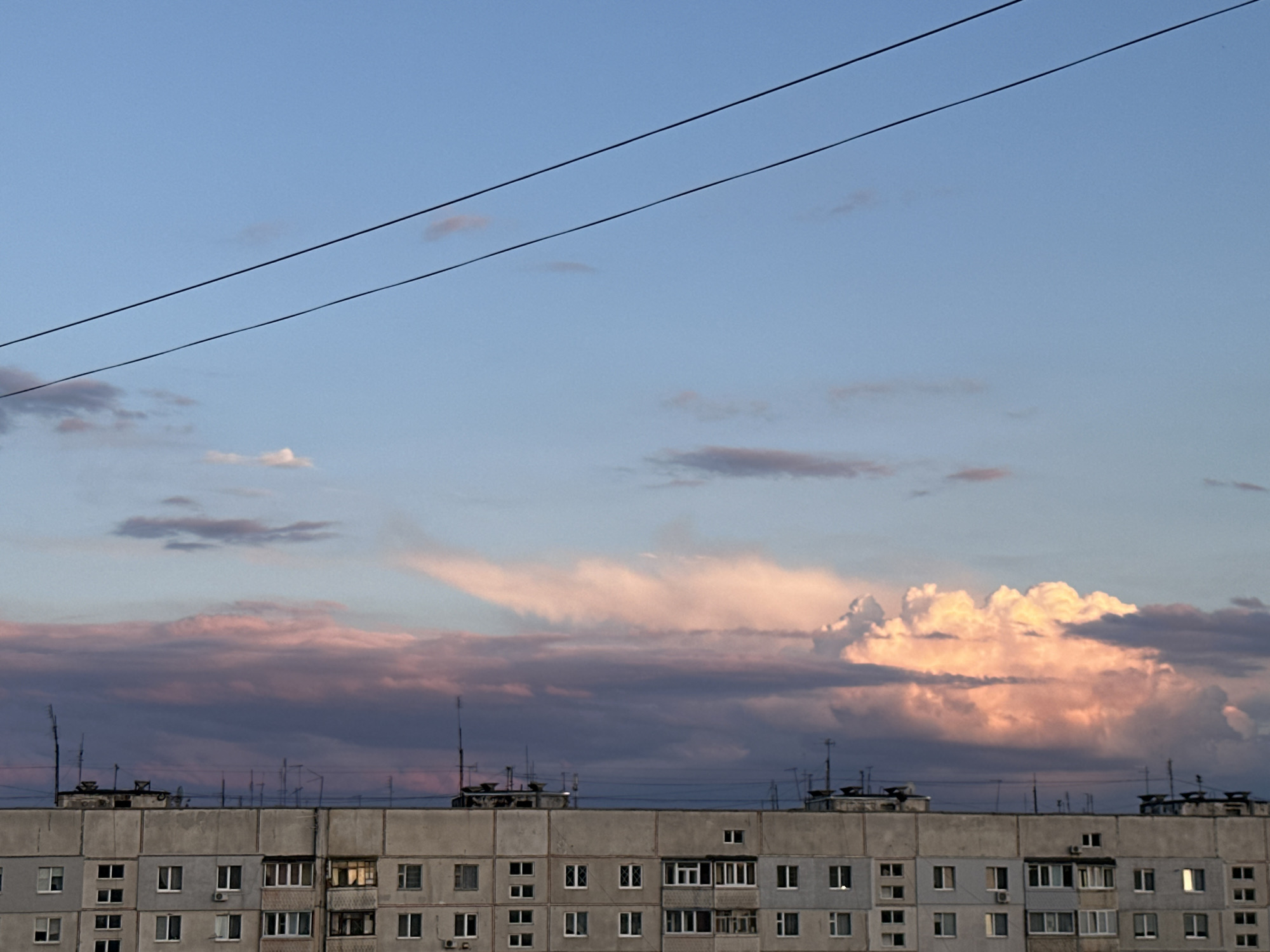 The image shows a long, gray, multi-story apartment building against a twilight sky. Two dark power lines stretch diagonally across the upper portion of the frame. The sky transitions from a light blue at the top to a soft pink and purple hue near the horizon, where clouds are illuminated by the setting sun. The apartment building, with its repeating pattern of windows, appears weathered and utilitarian. The rooftop of the building is lined with various antennas and structures. The scene is devoid of people and implies an urban, residential setting during the evening.