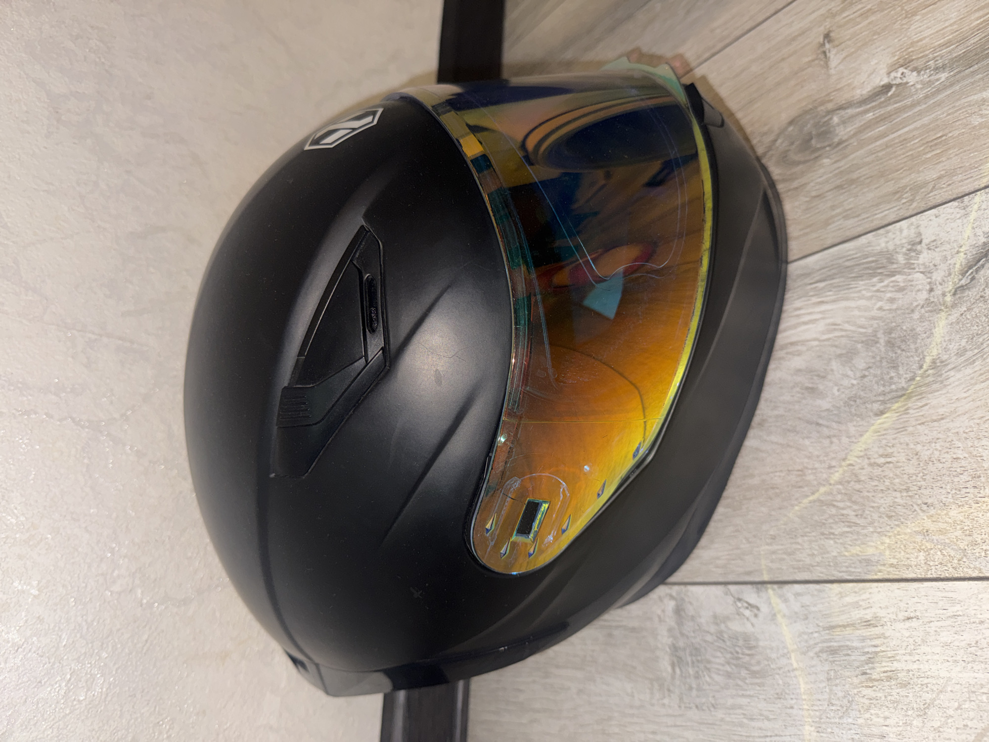A black motorcycle helmet with a reflective, multi-colored visor is positioned on a light-colored wooden floor. The helmet is the primary subject, shown from a side angle, slightly angled towards the viewer. The visor displays a rainbow-like iridescence, with visible scratches and some minor damage. The texture of the helmet appears to be matte. The background shows a textured wall and a dark horizontal line, possibly a baseboard or shelf. The floor has a wood grain pattern. The scene is indoors, and there are no people present. The lighting suggests an artificial light source. The location is the city of Bishkek, Kyrgyzstan.