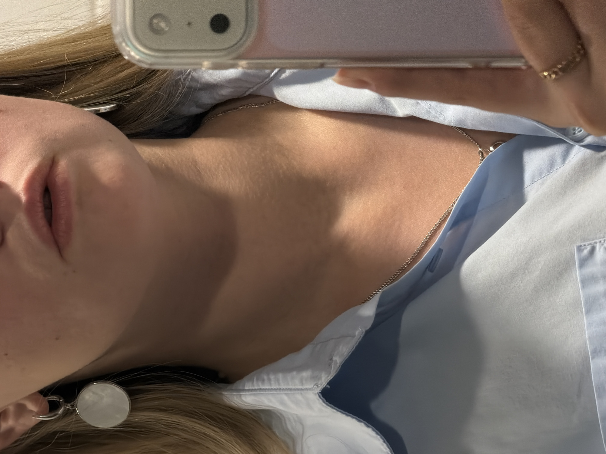 A close-up photograph depicts a fair-skinned person wearing a light blue button-down shirt and silver jewelry, including a necklace and an earring with a circular pendant. A mobile phone is positioned in the foreground, partially obscuring the view. The photo's orientation is slightly tilted. The location is not discernible from the image; there is no indication it was taken in Izhevsk, Russia.